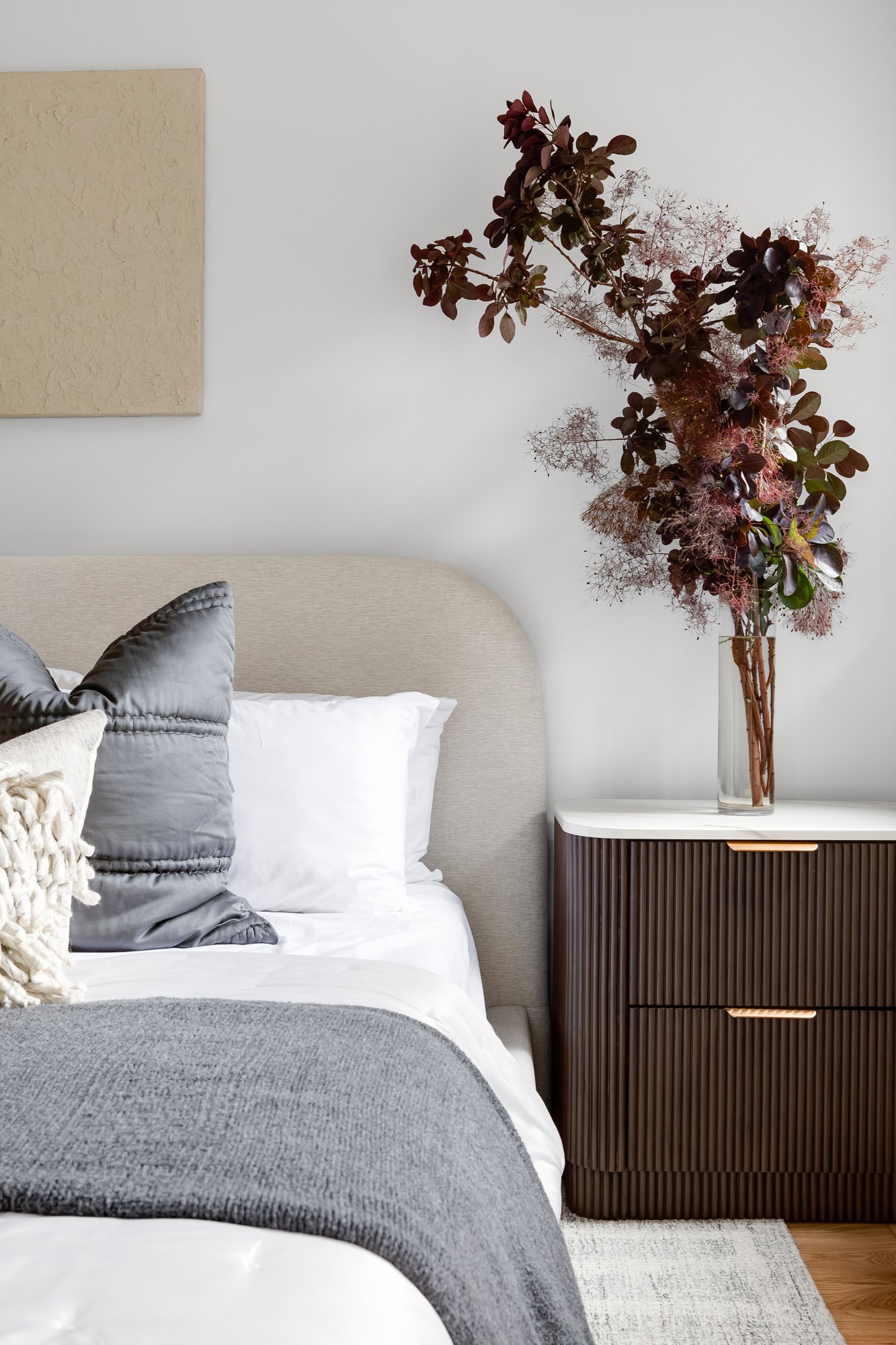 Primary bedroom detail in Castro San Francisco luxury TIC featuring linen upholstered headboard, layered gray bedding, fluted dark walnut nightstand with marble top and brass hardware, and dramatic dried smoke bush arrangement