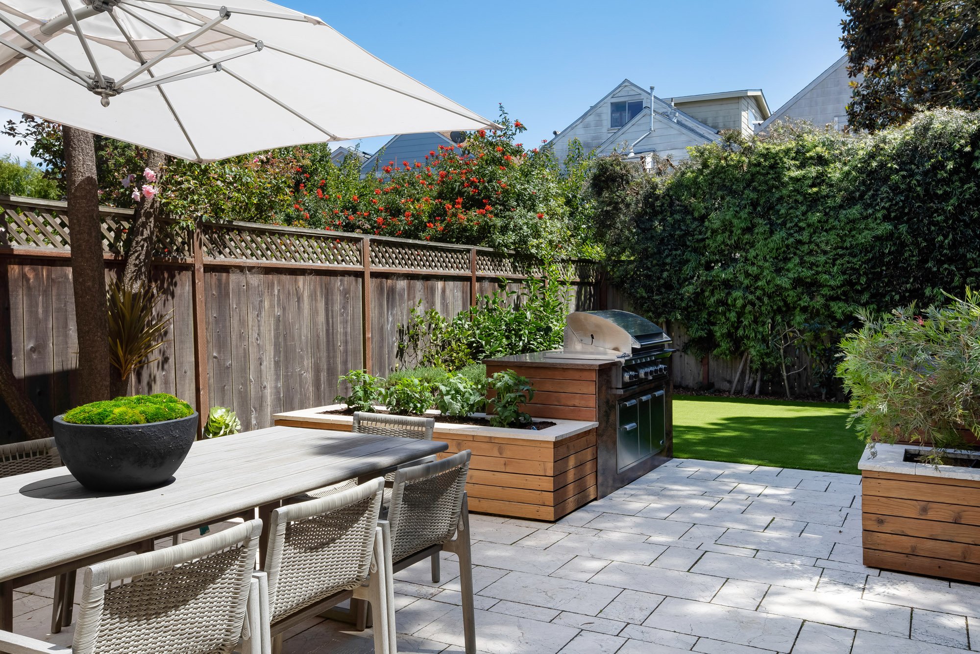 Sunny rear patio at 771 Elizabeth Street in Noe Valley with an outdoor dining table, built-in grill station, flowering roses along the fence, and a tidy artificial-turf lawn. 