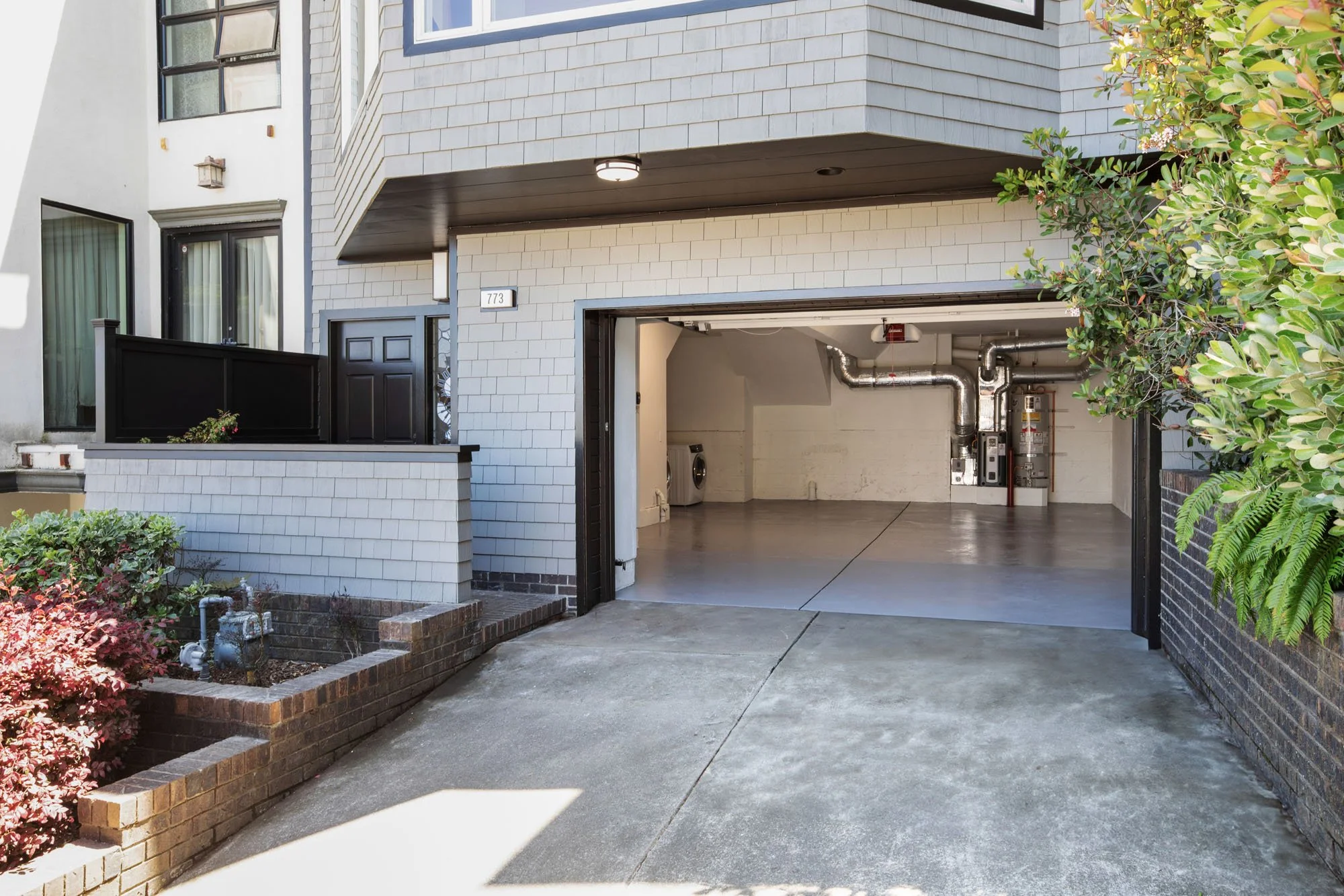 Exterior view of an open two-car garage with clean epoxy flooring and upgraded utility systems, showing the grey shingle siding and brick planter boxes.