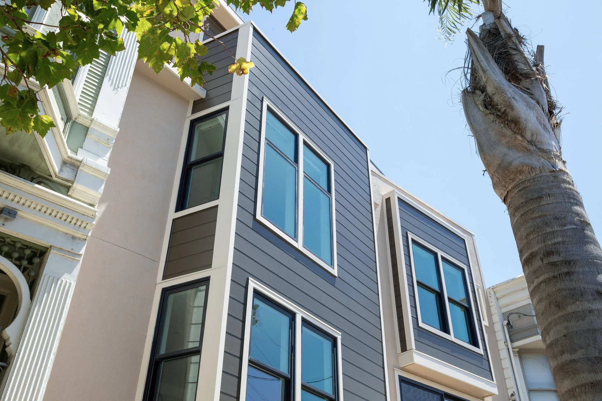 Architectural detail of a modern home facade showcasing dark grey siding, clean white trim, and large energy-efficient windows against a clear blue sky.