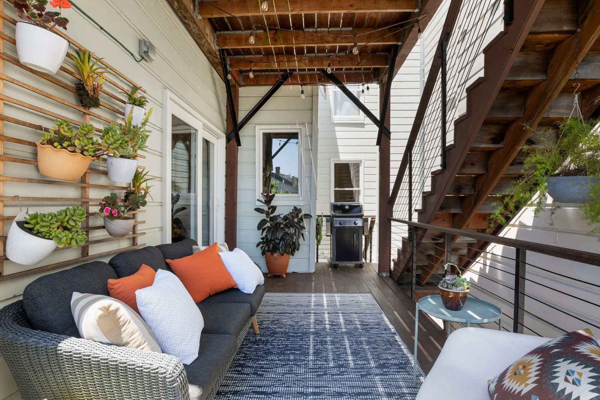 Private covered wood deck with a grey wicker sofa, blue patterned rug, and a vertical wall garden with potted succulents and flowers.