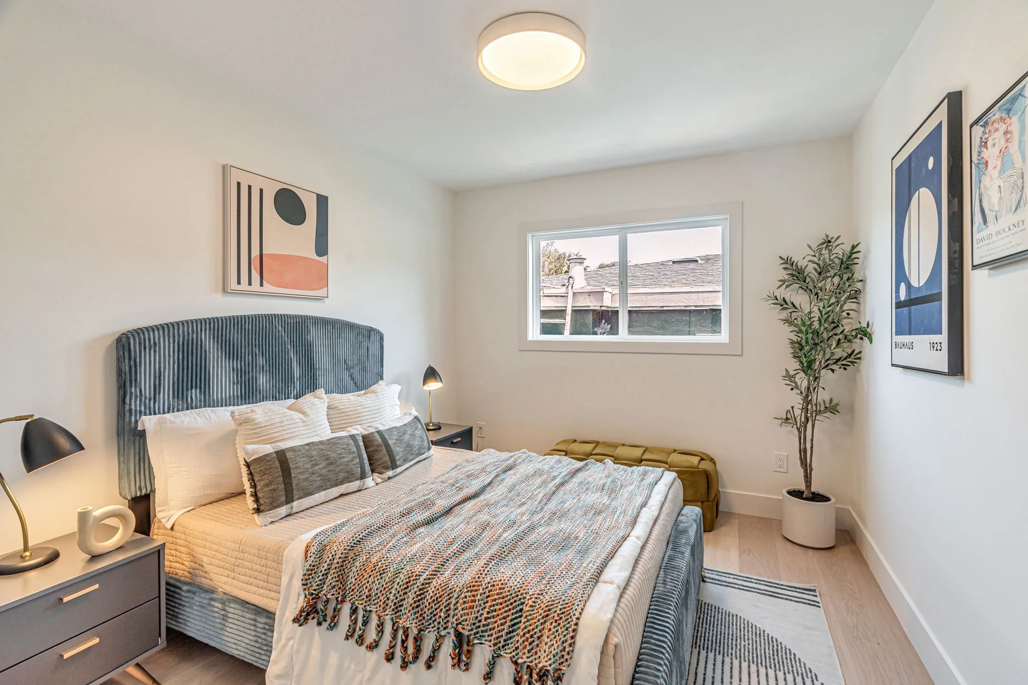 A full view of the third bedroom showing the blue-textured bed, a medium-sized window, and a mustard yellow velvet bench. The space is decorated with contemporary art prints and a potted olive tree.