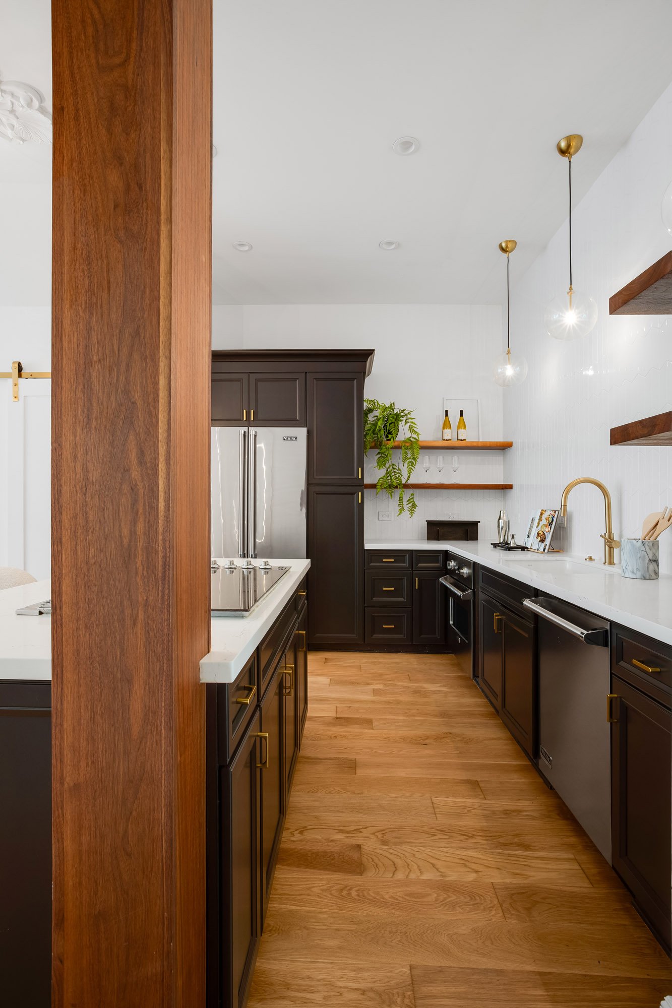 Luxury kitchen at Castro San Francisco TIC viewed past walnut structural beam, showing Viking stainless French door refrigerator, espresso Shaker cabinets with brass pulls, walnut open shelving, globe pendant lights, and white quartz countertops