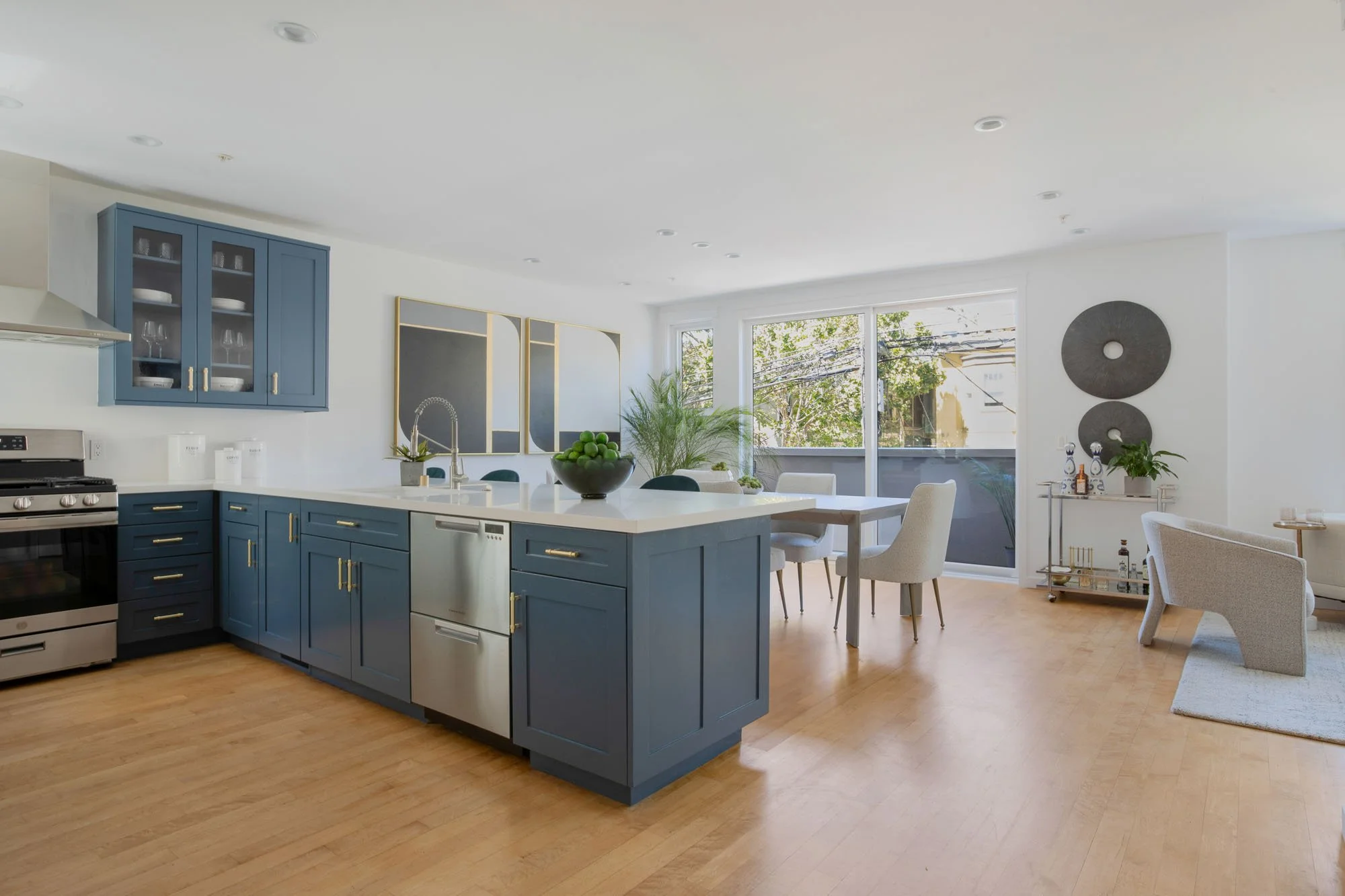 A wide-angle view of a modern open-concept kitchen and living space. It features slate blue shaker-style cabinetry with gold hardware, white quartz countertops, and a large island. In the background, a light-filled dining area sits in front of a slid