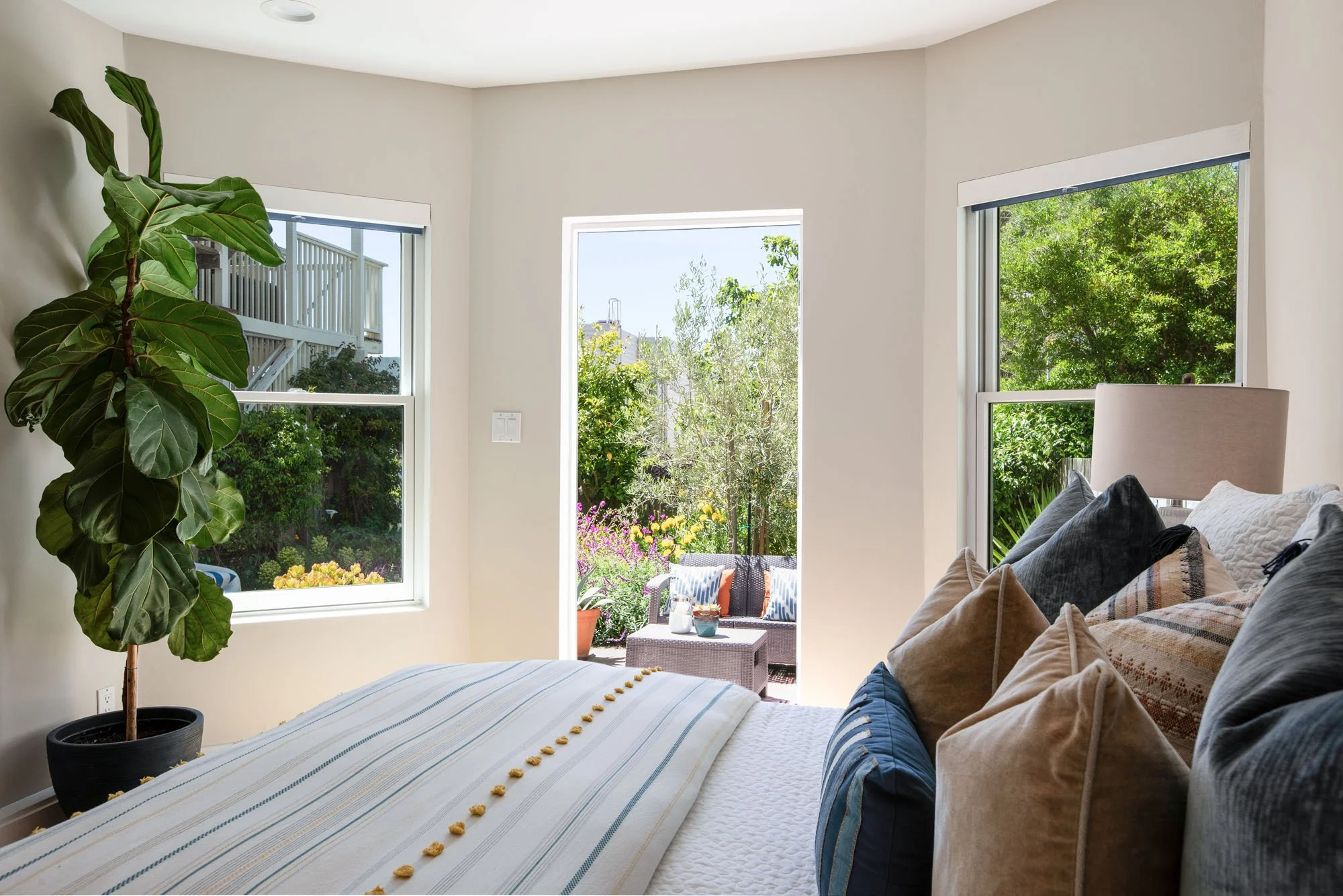 View from a sunlit bedroom through an open door to a lush garden patio with outdoor seating and vibrant purple flowers.