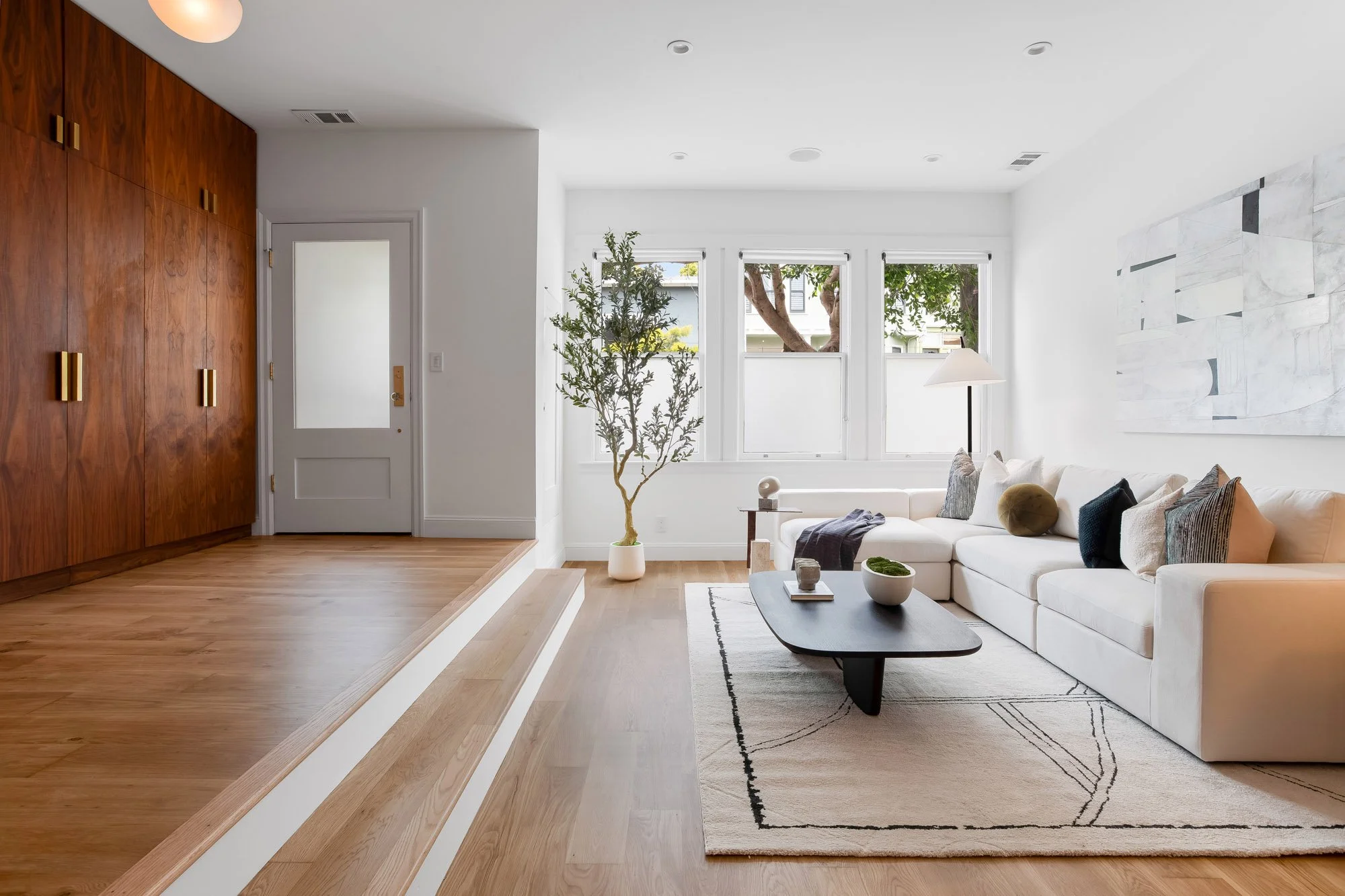 Sunlit living room in 4545 18th Street San Francisco TIC featuring raised sitting area with custom walnut built-ins, cream sectional sofa, black organic coffee table, Moroccan-style area rug, and triple street-facing windows