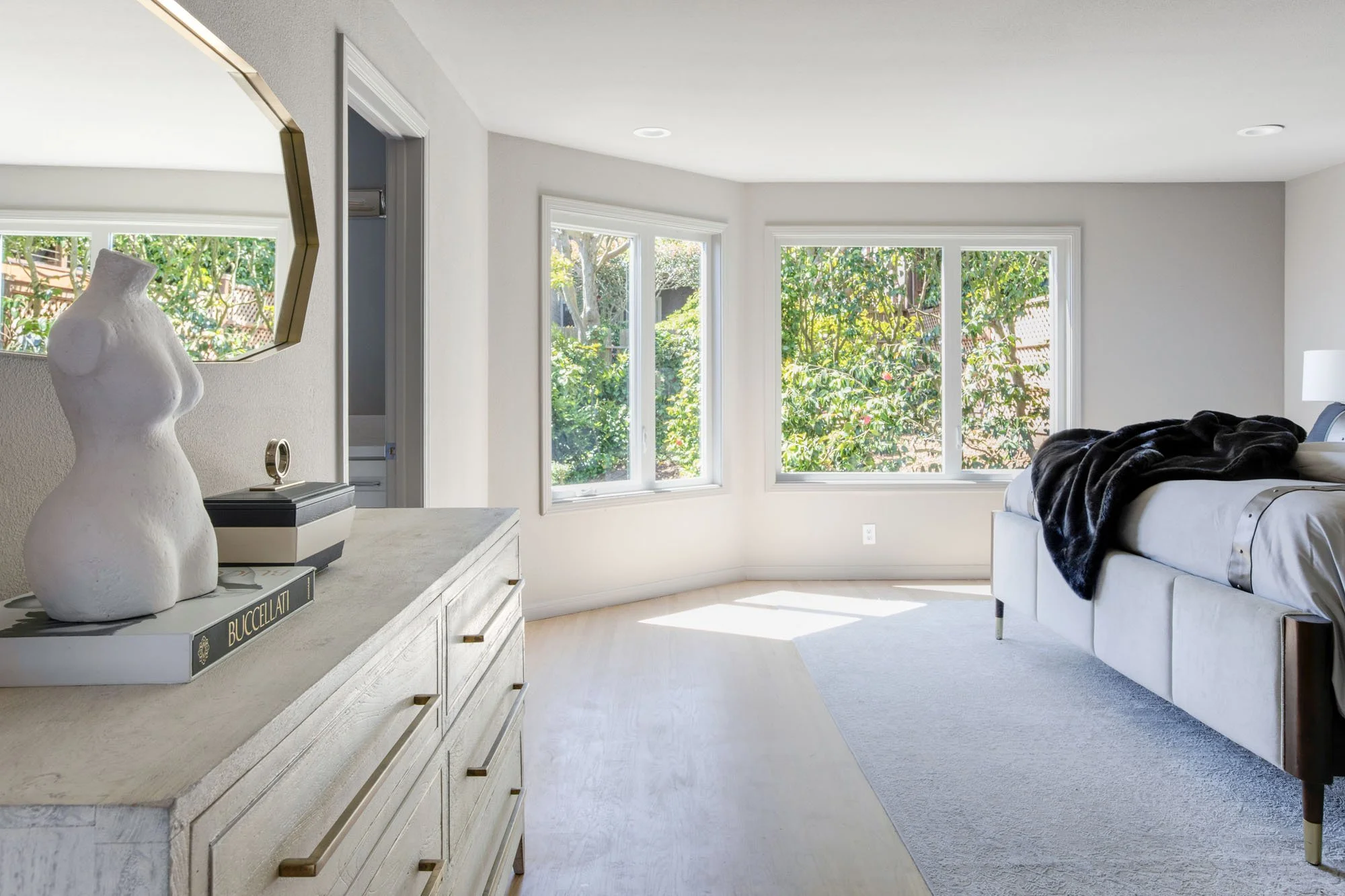 Primary bedroom suite featuring large bay windows, light oak hardwood floors, and a designer dresser with a minimalist bust sculpture and gold-framed mirror.