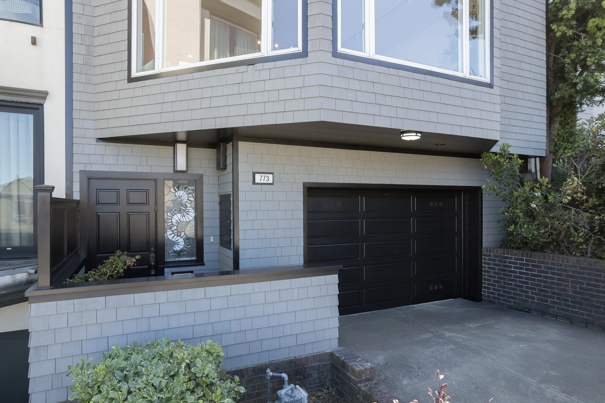 Full street-level view of a contemporary hillside residence in San Francisco, showcasing expansive bay windows, professional landscaping, and modern architectural grey wood shingles.