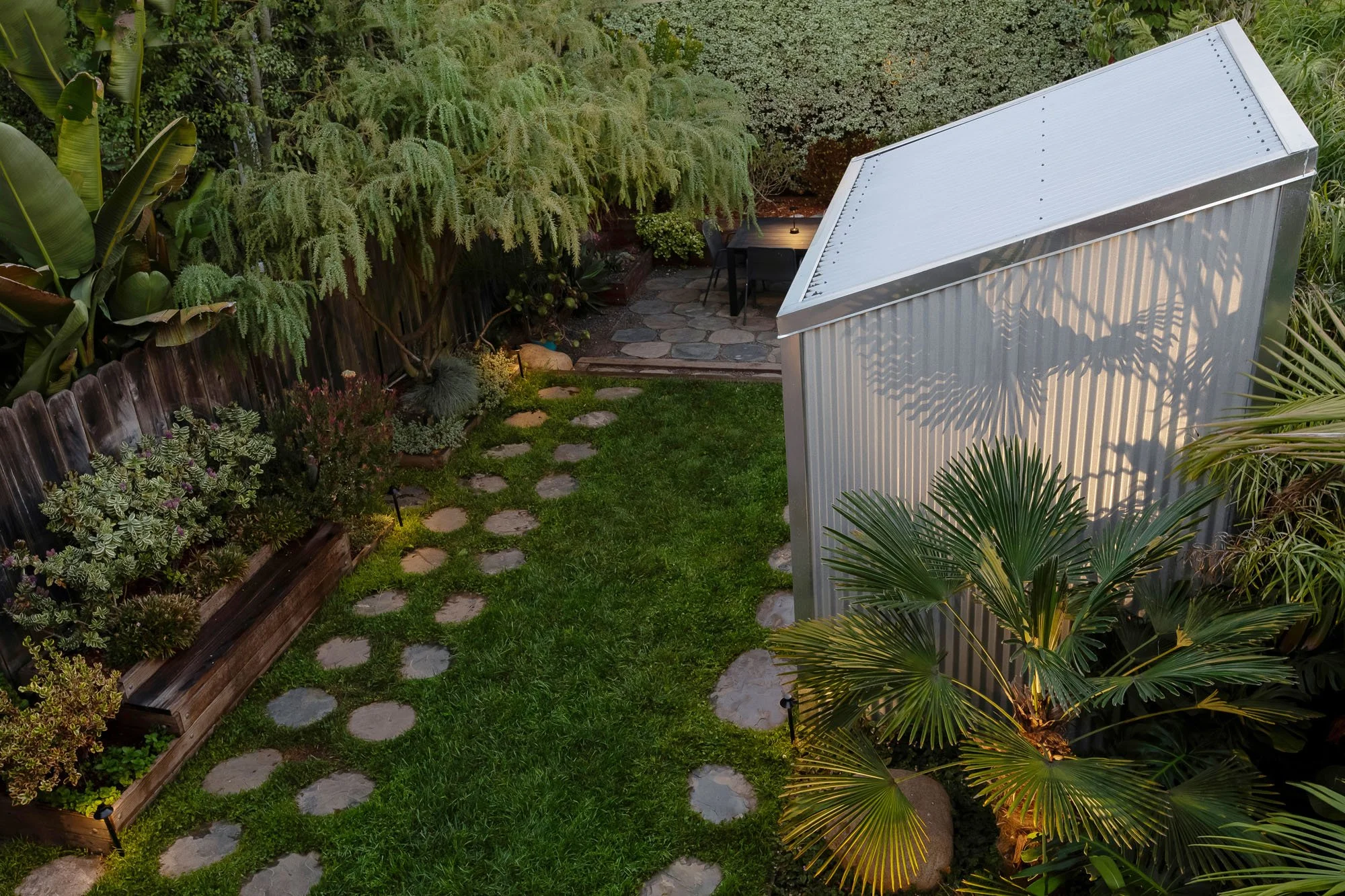 High-angle evening view of the backyard at 246 Detroit Street, SF, featuring a modern silver corrugated metal garden studio, a circular stone paver path through a green lawn, and warm landscape lighting among lush tropical plants.
