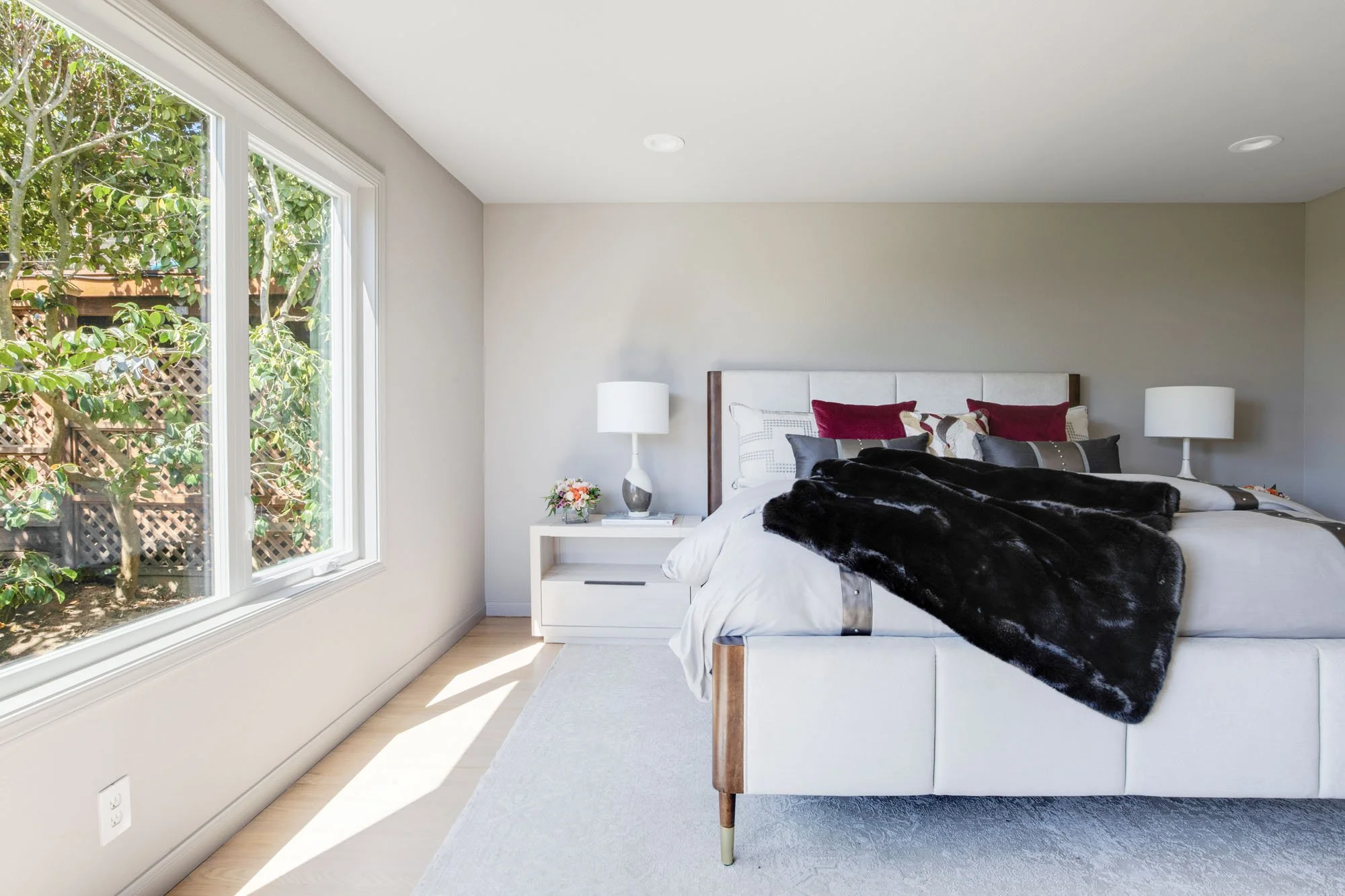 Spacious primary bedroom with a contemporary white upholstered bed, dark faux-fur throw, and large windows offering views of the private garden and greenery.
