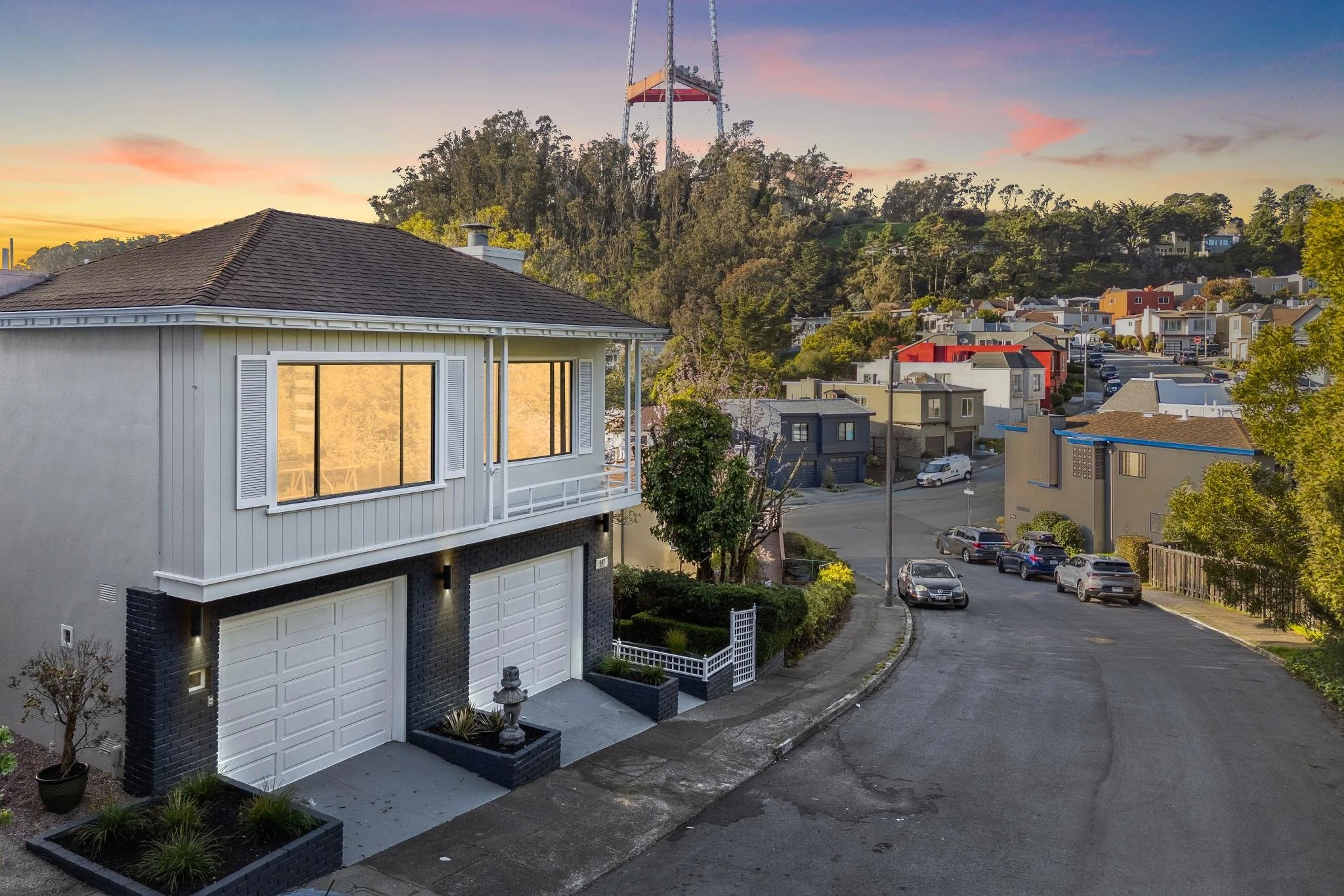 Aerial view of a modern two-story home at 147 Gladeview Way, San Francisco, featuring two white garage doors and a grey brick facade with Sutro Tower in the background at sunset.