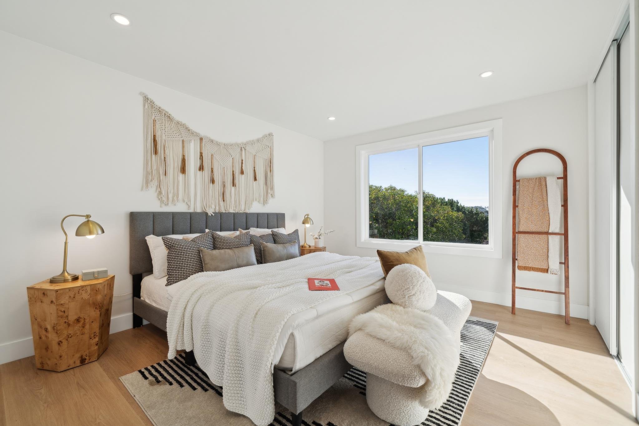 Sophisticated primary bedroom featuring a grey upholstered bed, macramé wall hanging, designer gold lamps, and a large window with views of the San Francisco hills.