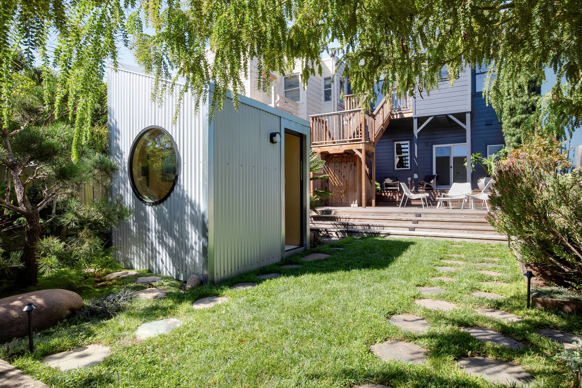 A wide-angle view of a tiered San Francisco backyard under the shade of a large tree, showcasing the contrast between the metal studio and the navy blue residential exterior.