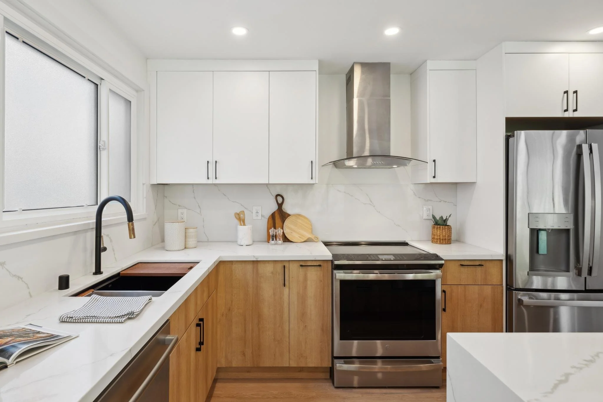 Modern kitchen view featuring a stainless steel range hood, white and wood cabinetry, and a large window providing ample natural light over the marble countertops.