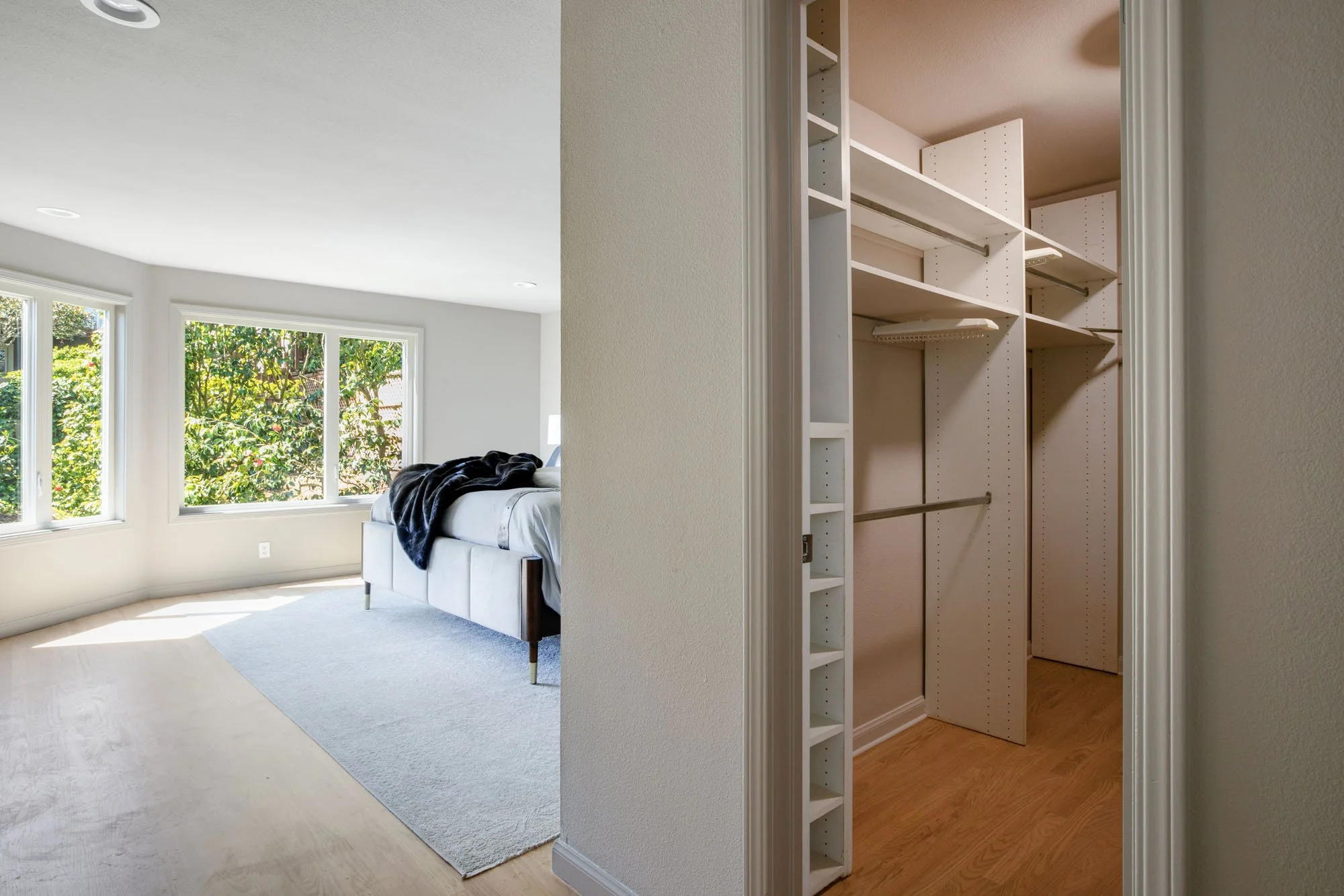 View of a walk-in primary closet with a white custom organization system, leading into a bright, sun-drenched bedroom with large garden-view windows.