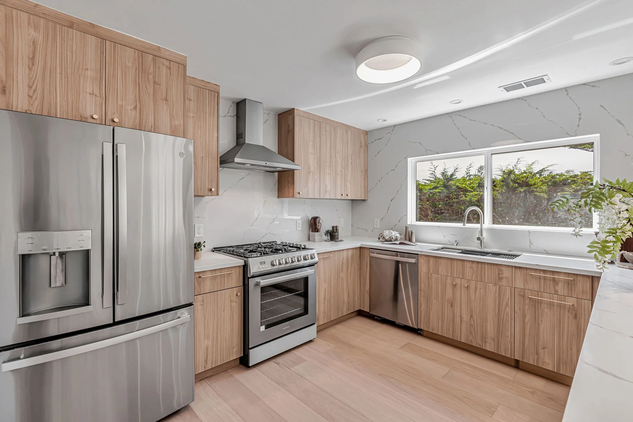 A bright, contemporary kitchen featuring light wood cabinetry with minimalist gold hardware and white marble-look quartz countertops. The space is equipped with high-end stainless steel appliances, including a French-door refrigerator, a gas range wi