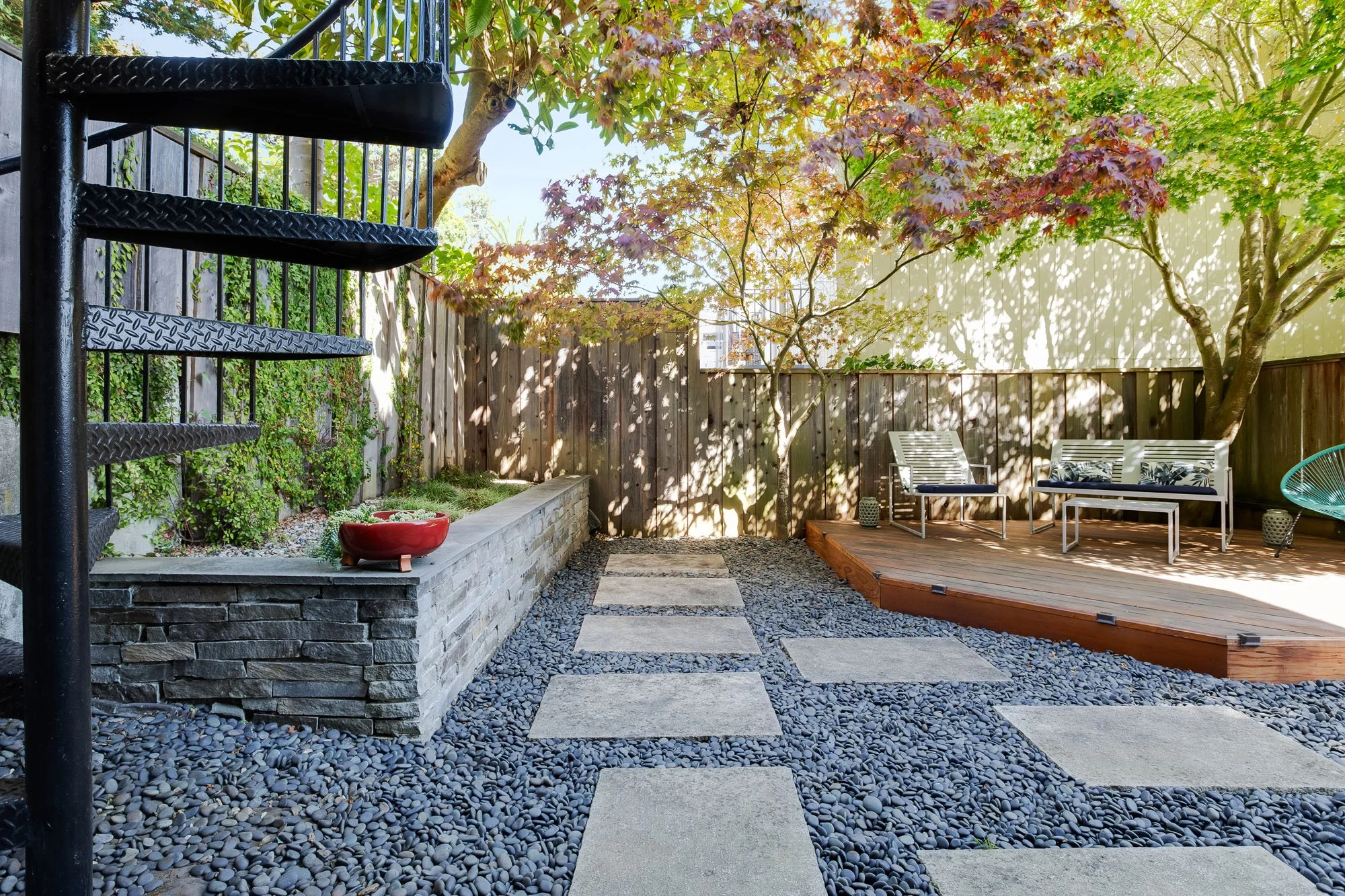A professionally landscaped San Francisco backyard featuring a black metal spiral staircase, a stone retaining wall, and a gray pebble ground cover with square concrete pavers.