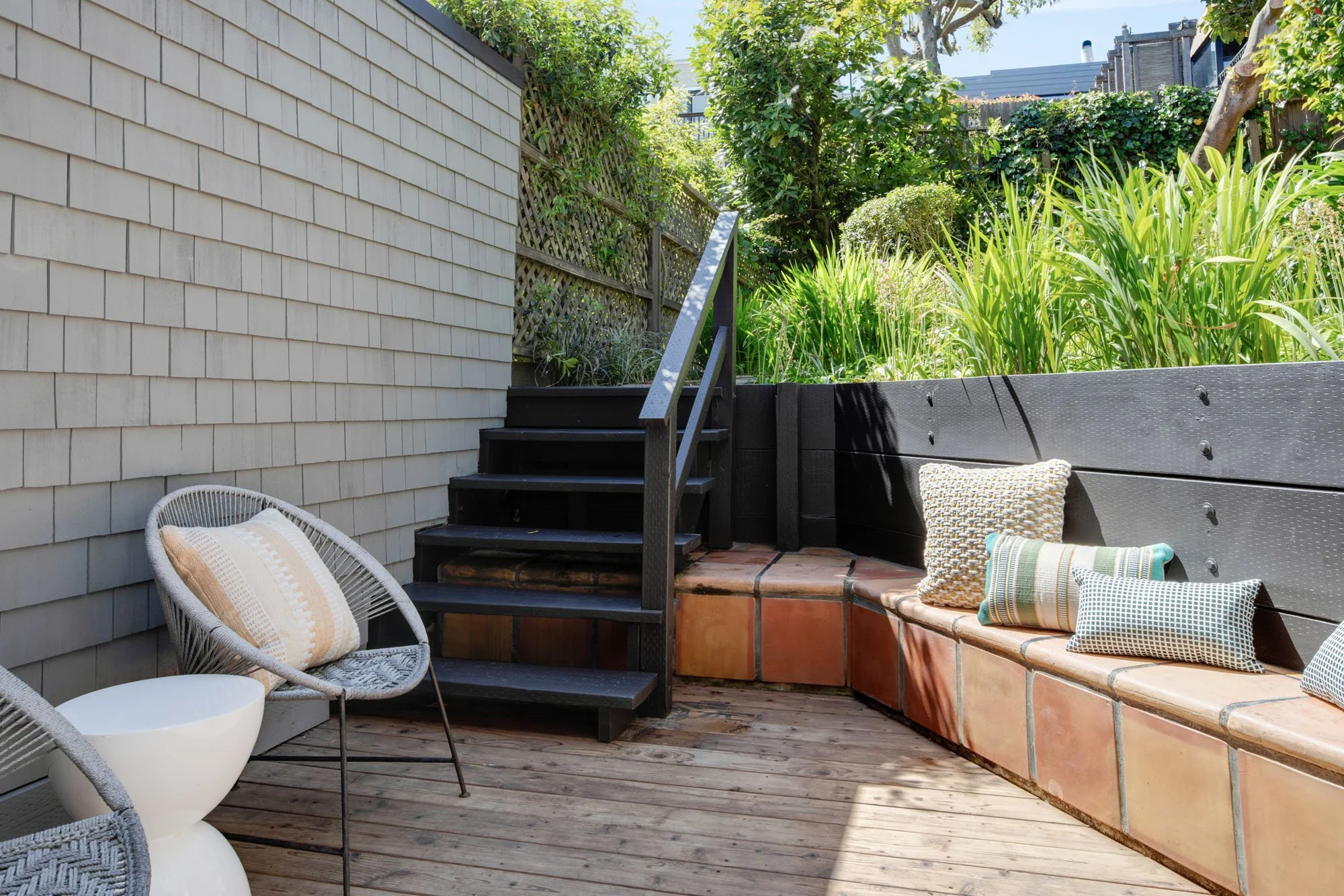 Private outdoor terrace with a tiered wooden deck, built-in terracotta-tiled seating, and modern grey wicker chairs surrounded by lush green garden landscaping.