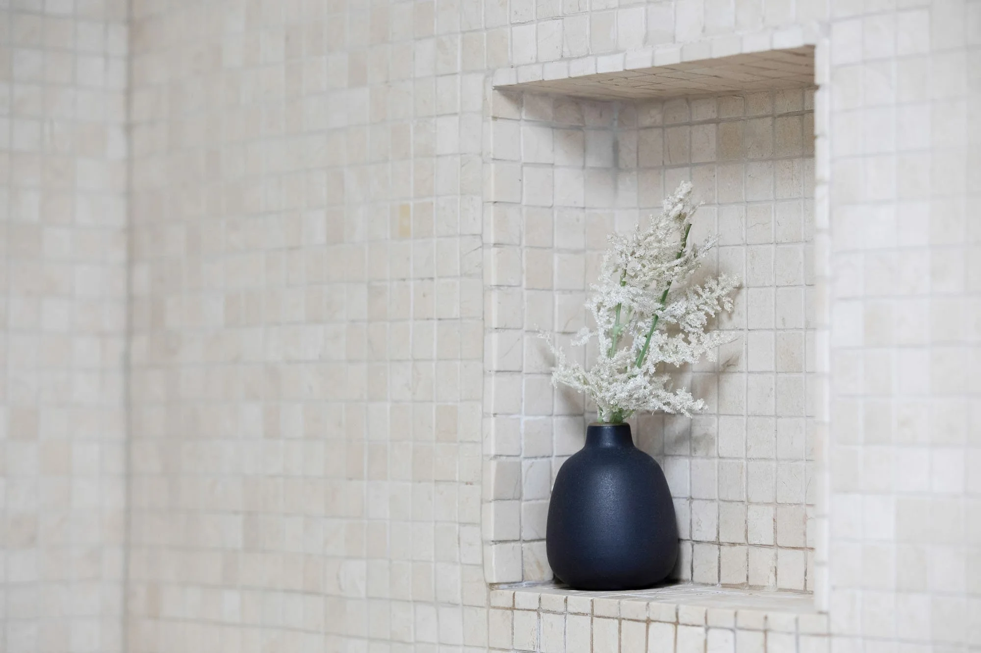 Interior design detail showing a black ceramic vase with dried white florals tucked into a built-in shower niche with neutral mosaic tiling.