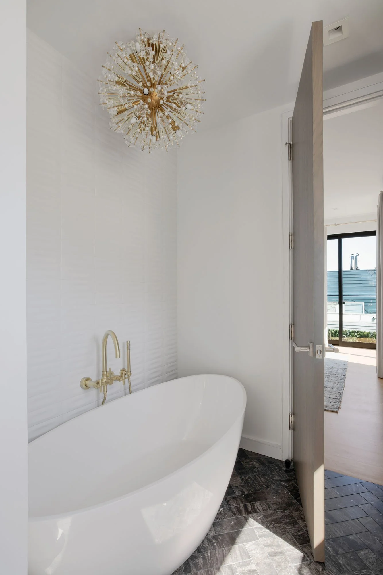 Luxury jack-and-jill bathroom at 1647 Sanchez featuring a freestanding soaking tub, brushed gold fixtures, and a sunburst crystal chandelier over dark herringbone tile.