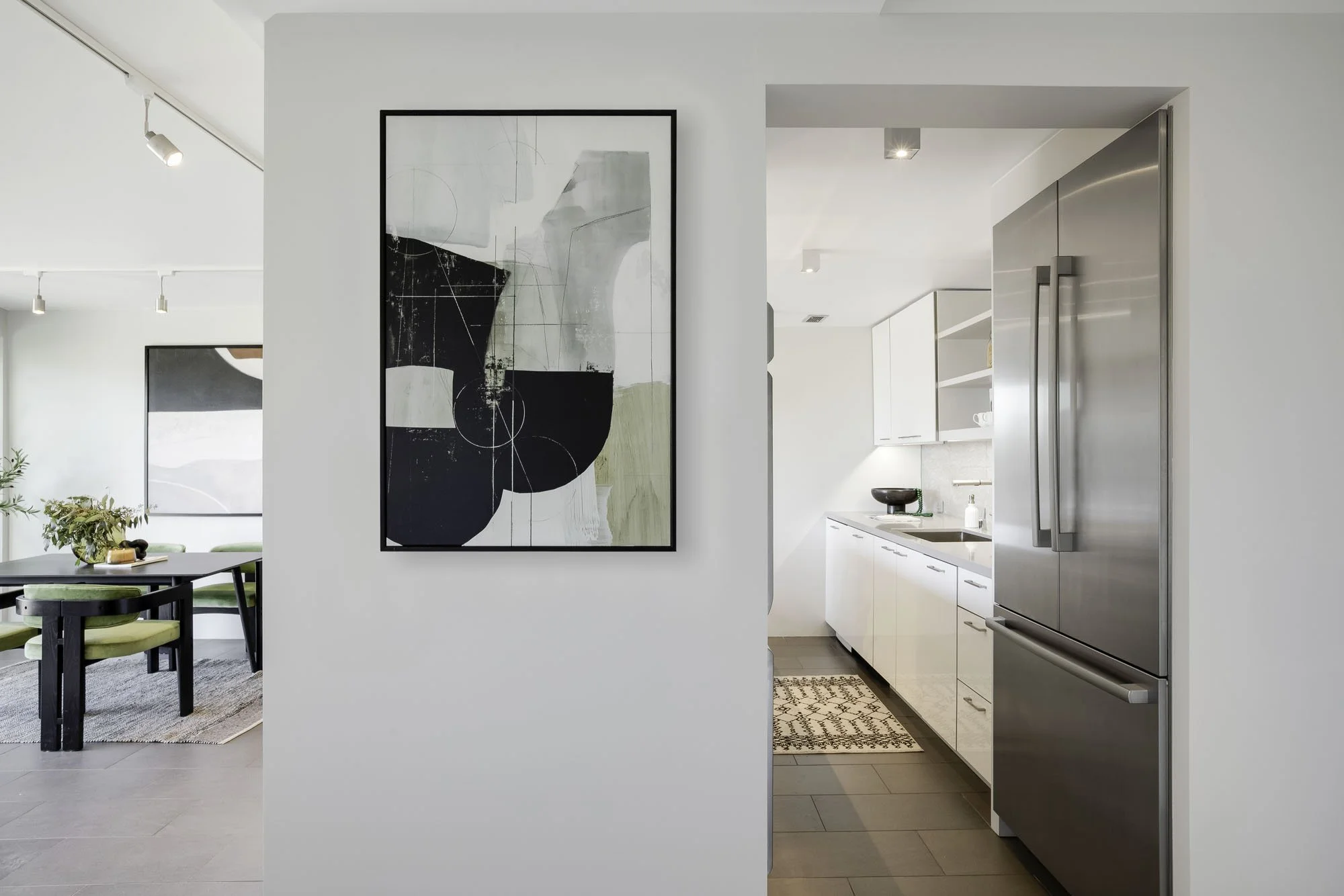 An interior view looking from a living/dining space through a doorway into a modern kitchen, with a central white column in the foreground. On the column hangs a large, framed abstract geometric painting. To the right, through the opening, the renova