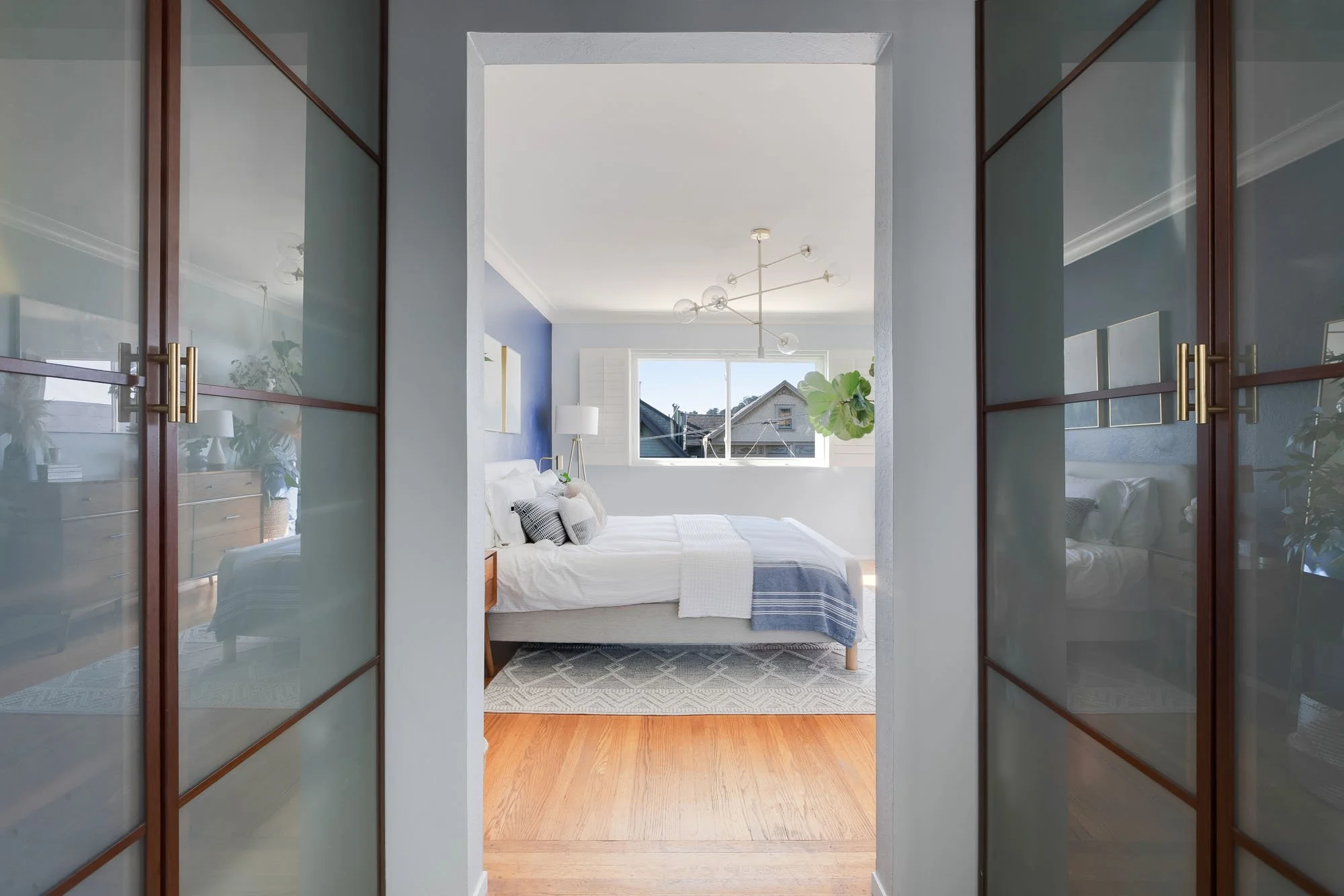 Looking through a dressing area with frosted glass sliding closet doors into the sunlit primary bedroom.