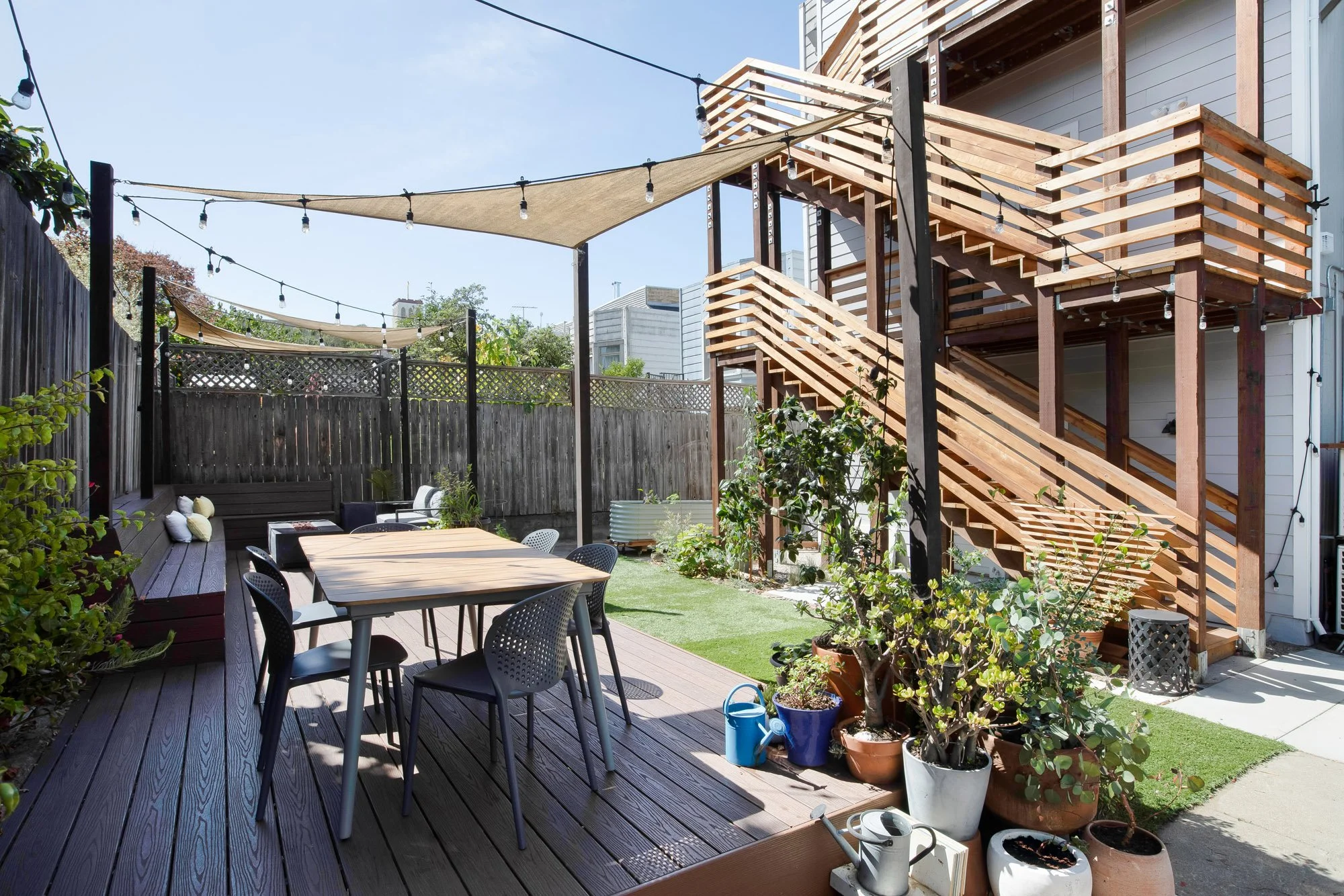 Shared rear yard with a sail-shade canopy, string lights, and built-in bench seating below the building's exterior stairs
