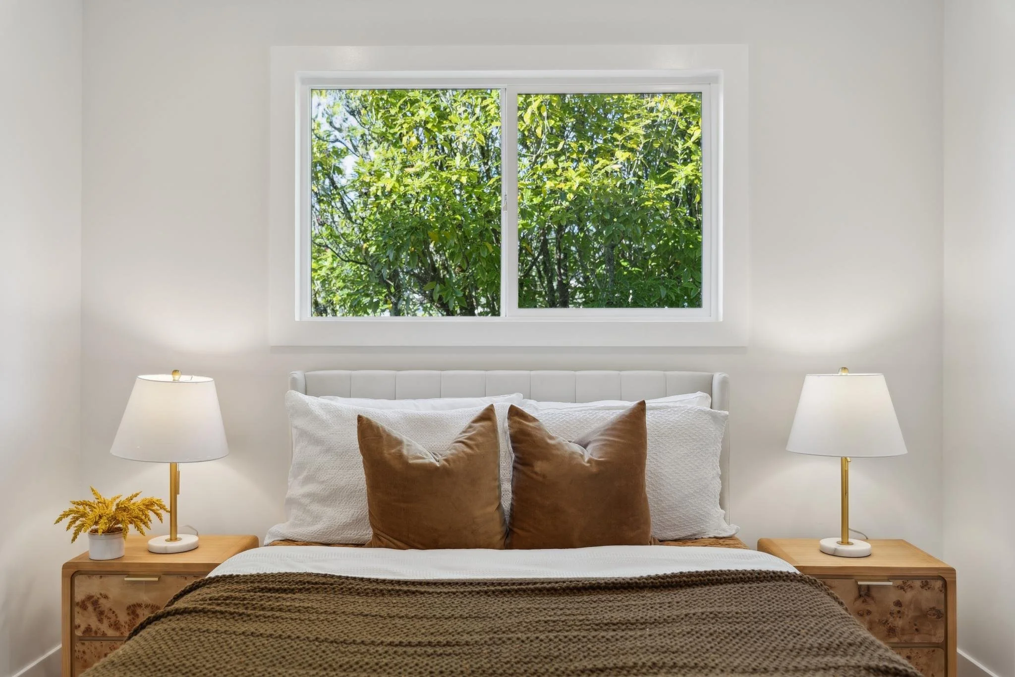Centered view of a cozy bedroom setup with symmetrical burl wood nightstands, gold lamps, and a white-framed window.