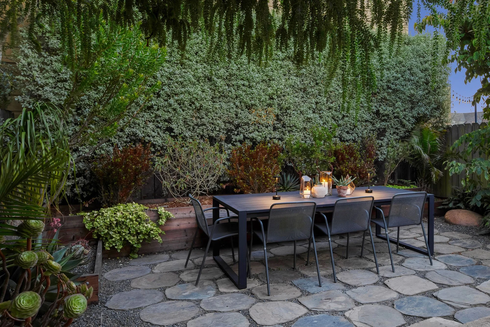 Secluded outdoor dining patio at 246 Detroit Street, SF, featuring a modern black metal table and mesh chairs on a flagstone surface, illuminated by cordless lanterns and surrounded by a lush, private garden hedge.