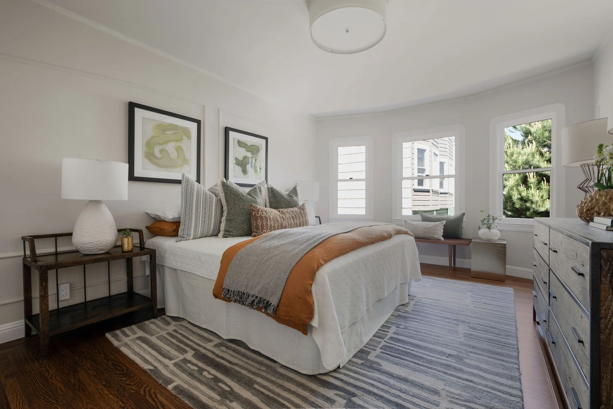 Second bedroom staged with earthy tones, a rustic wood dresser, and large windows, showcasing the home's versatile and airy floor plan.