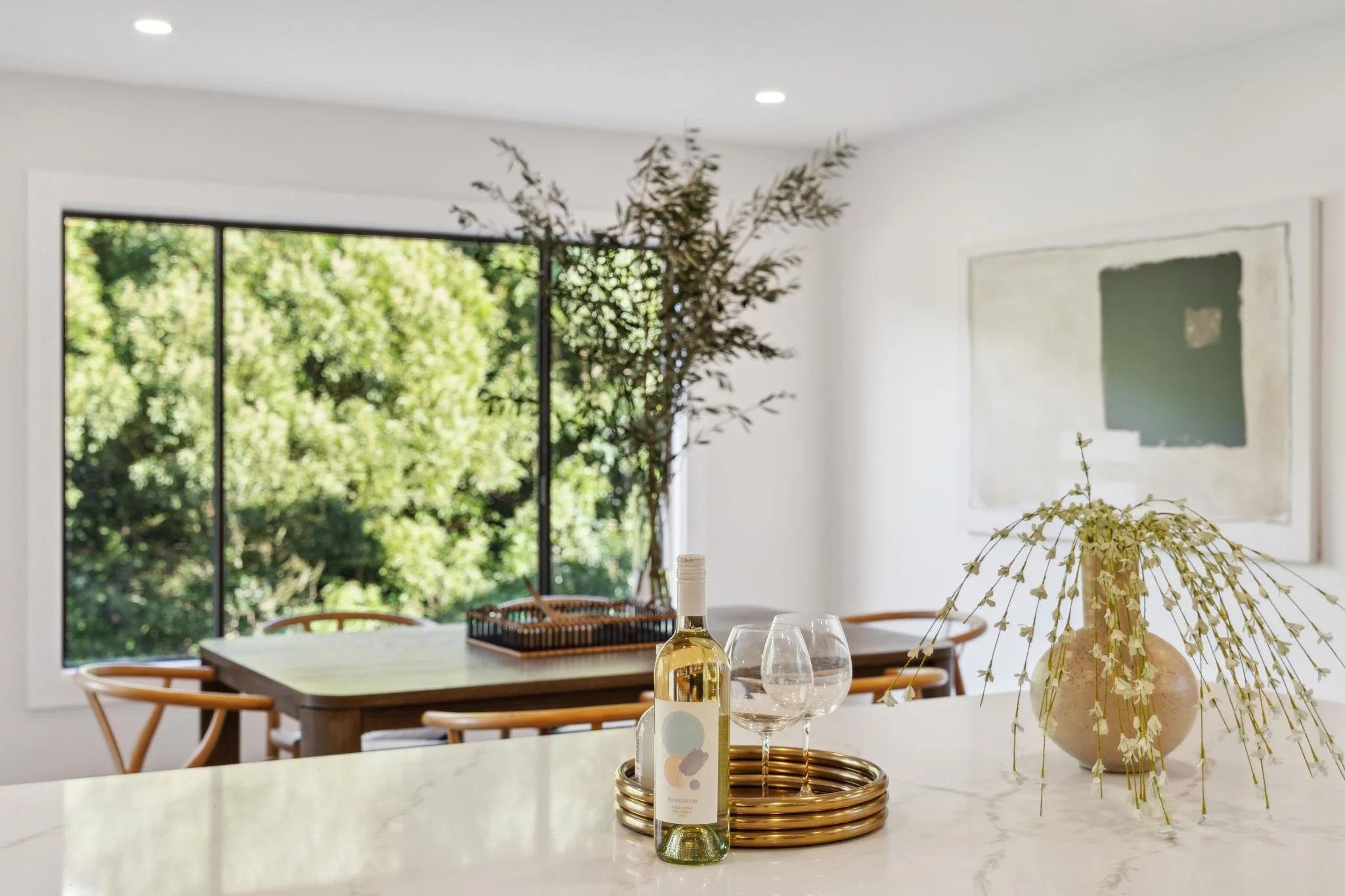 Close-up of a white quartz kitchen island with wine glasses and a bottle, looking out toward a dining area and a large window with tree views.