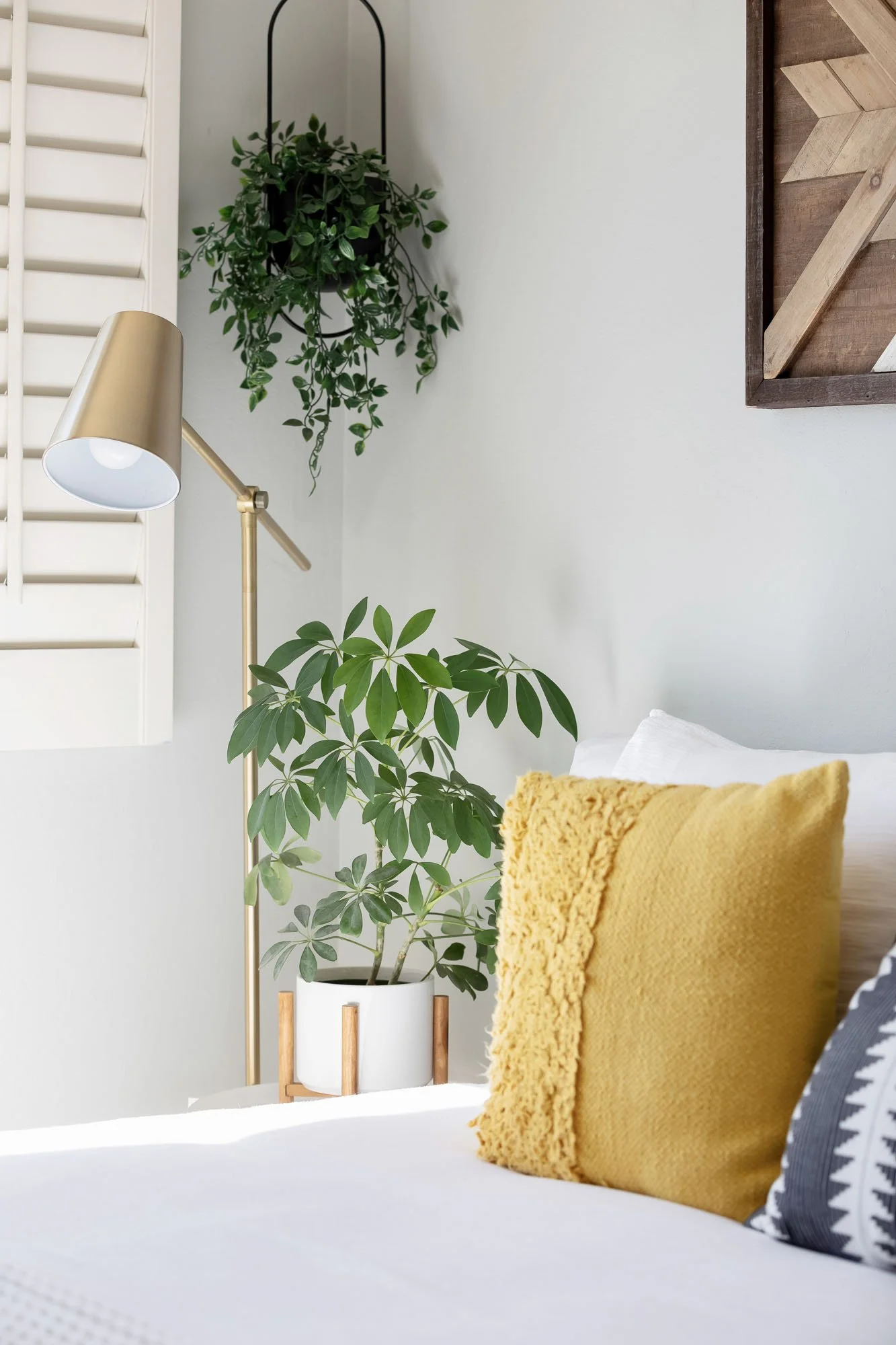 A cozy bedroom corner detail with a yellow textured pillow, a gold floor lamp, and a healthy Schefflera houseplant in a white ceramic pot.