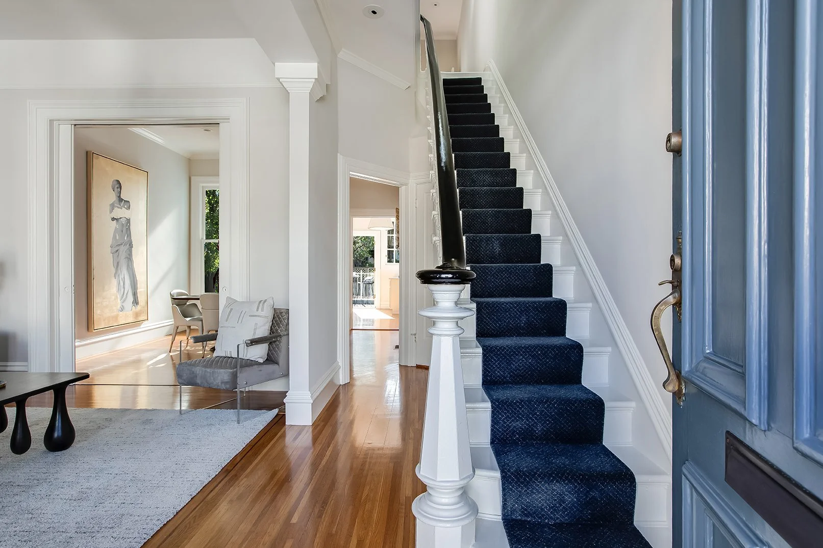 Bright foyer and grand staircase of a renovated luxury Victorian home, showcasing original hardwood floors and a modern navy carpet runner.