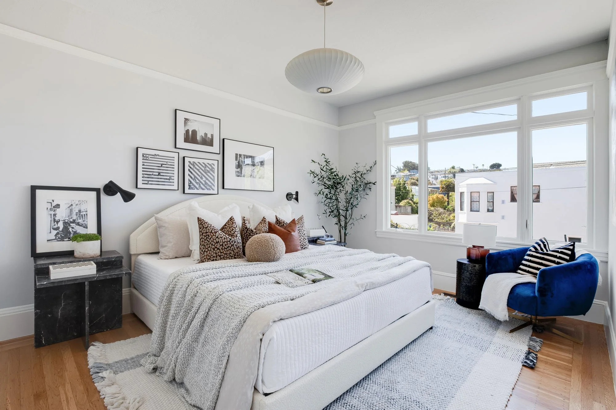 Bright bedroom at 246 Detroit St with white walls, a gallery wall of black and white photography, a marble side table, and a deep blue velvet accent chair by the window.