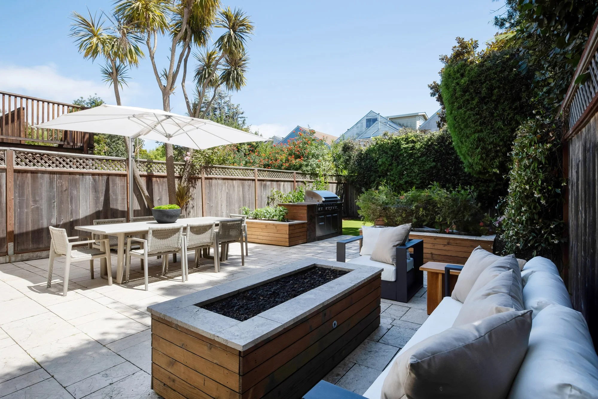 Flat, sunny backyard at 771 Elizabeth Street with a stone-paved patio, built-in gas grill, linear fire feature, raised garden beds, and shaded dining and lounge seating.