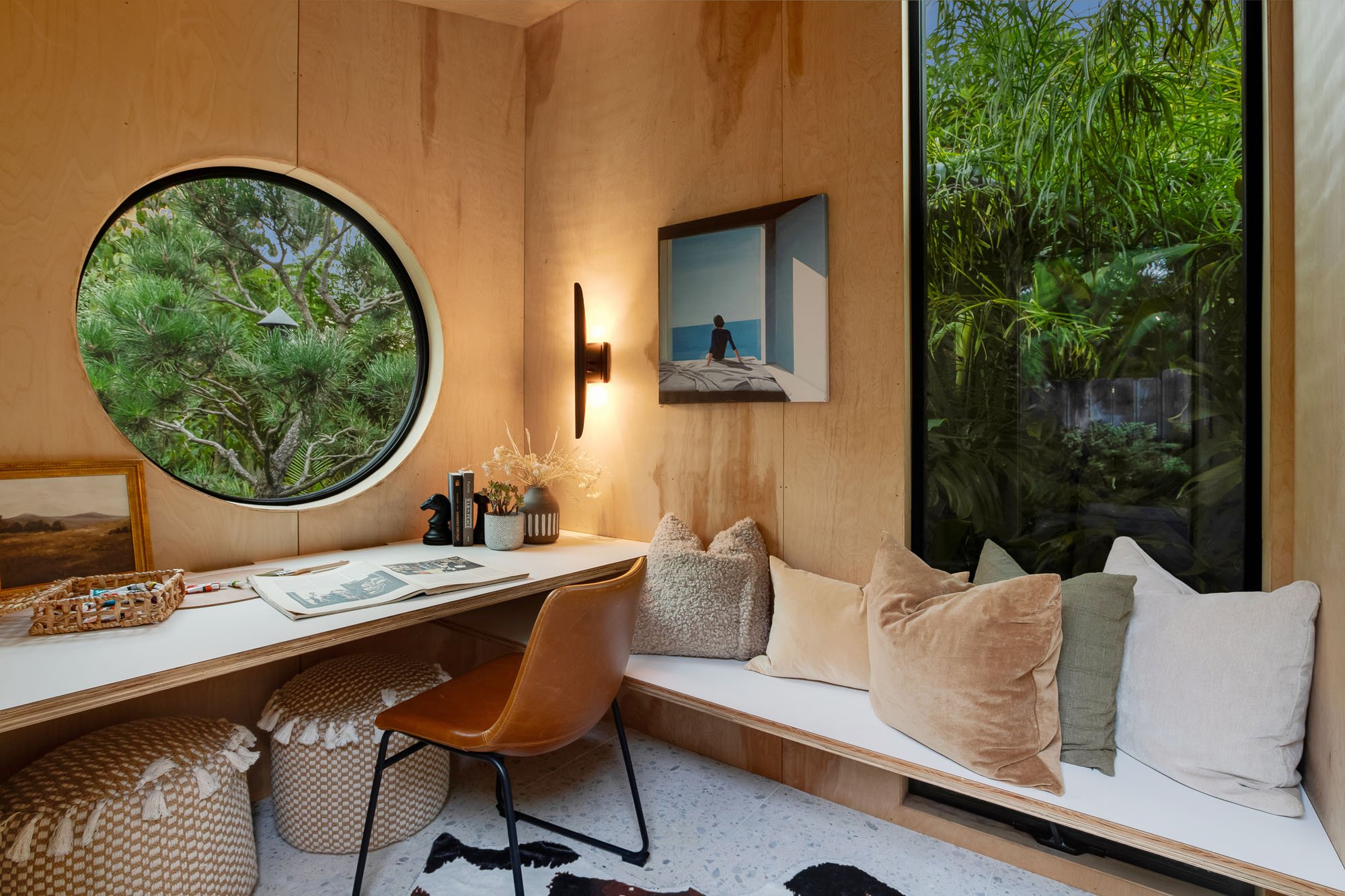 Modern garden studio interior at 246 Detroit Street, SF, featuring minimalist plywood walls, a large circular porthole window, a built-in desk with a leather chair, and a cozy reading nook with plush pillows and warm sconce lighting