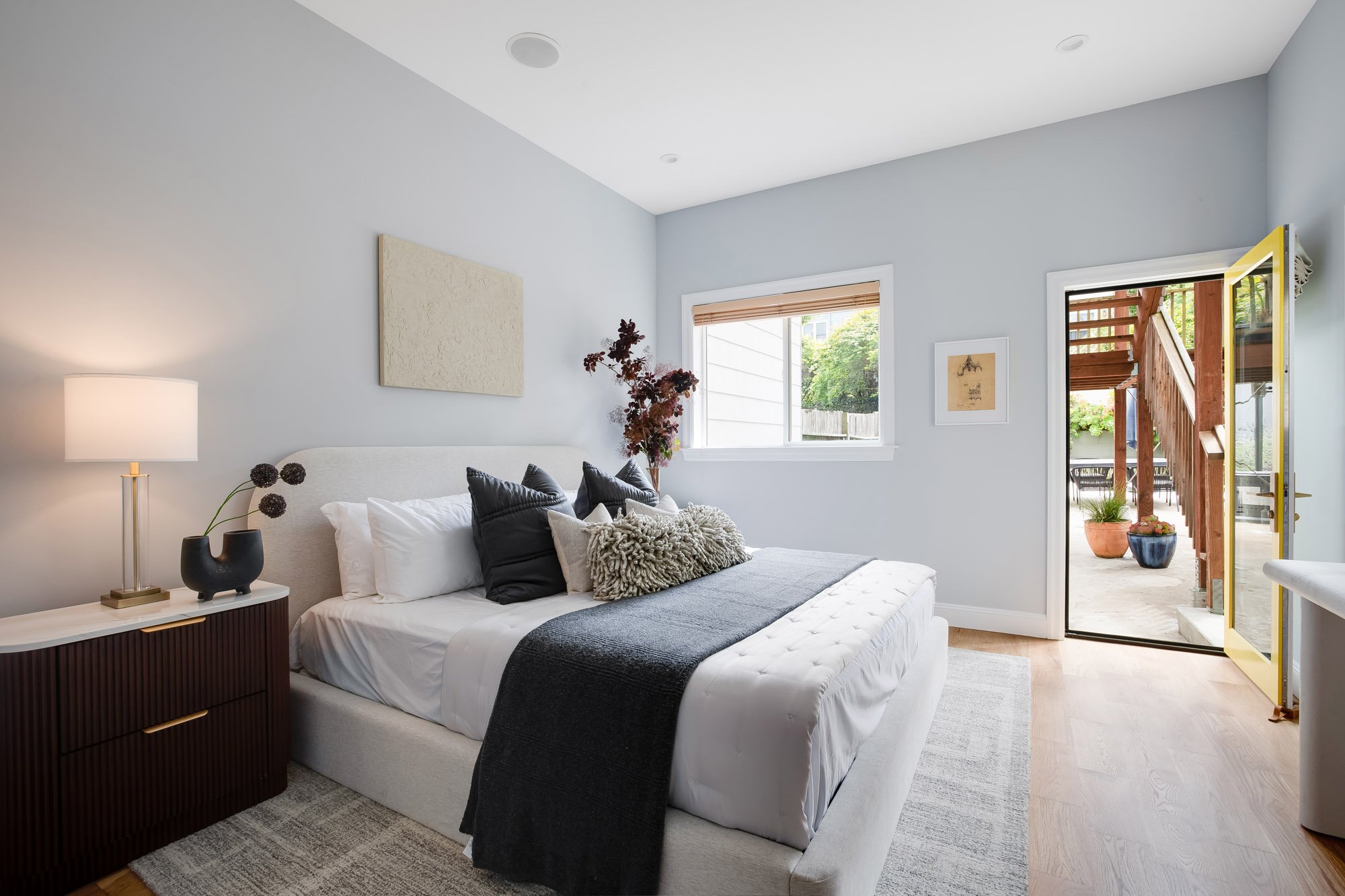 Primary bedroom in Castro San Francisco luxury TIC with upholstered platform bed, fluted dark walnut nightstand, brass table lamp, white oak floors, garden view window, and yellow-framed door opening to private rear deck and outdoor staircase