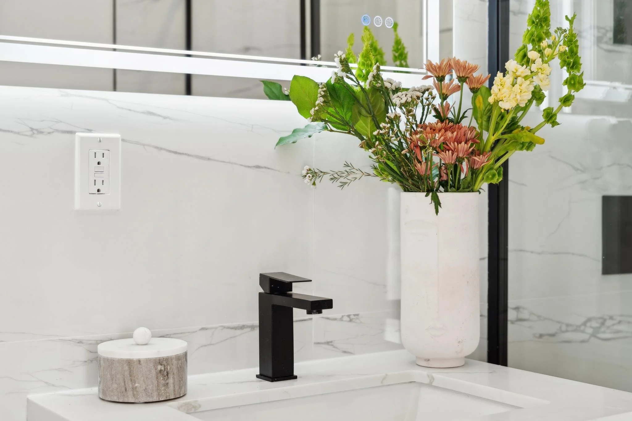 Close-up of a bathroom vanity with a white quartz countertop, a sleek matte black faucet, and a decorative floral arrangement.