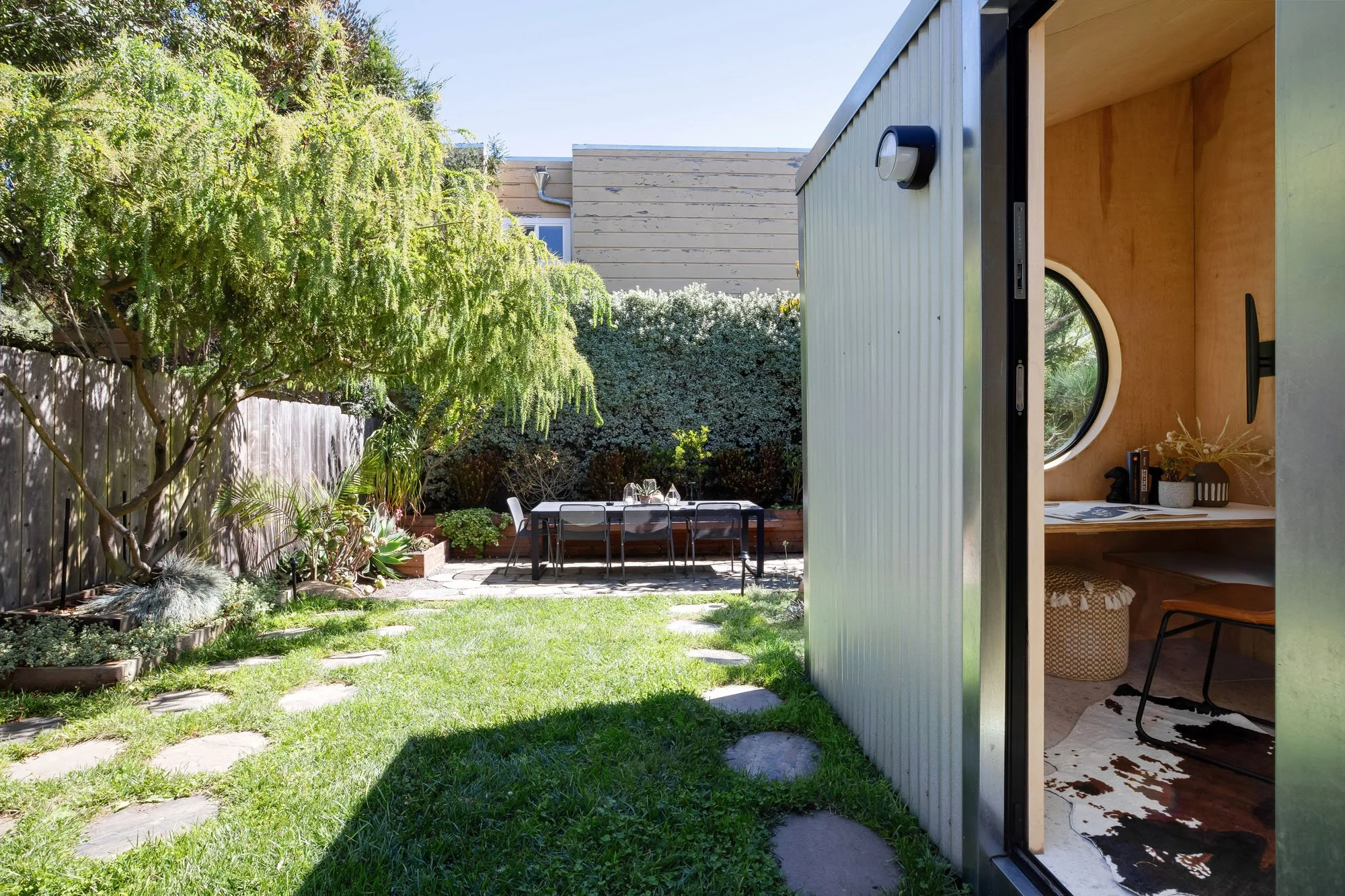 Looking through the open door of the corrugated metal garden studio to reveal a minimalist interior with plywood walls, a built-in desk, and a cowhide rug.