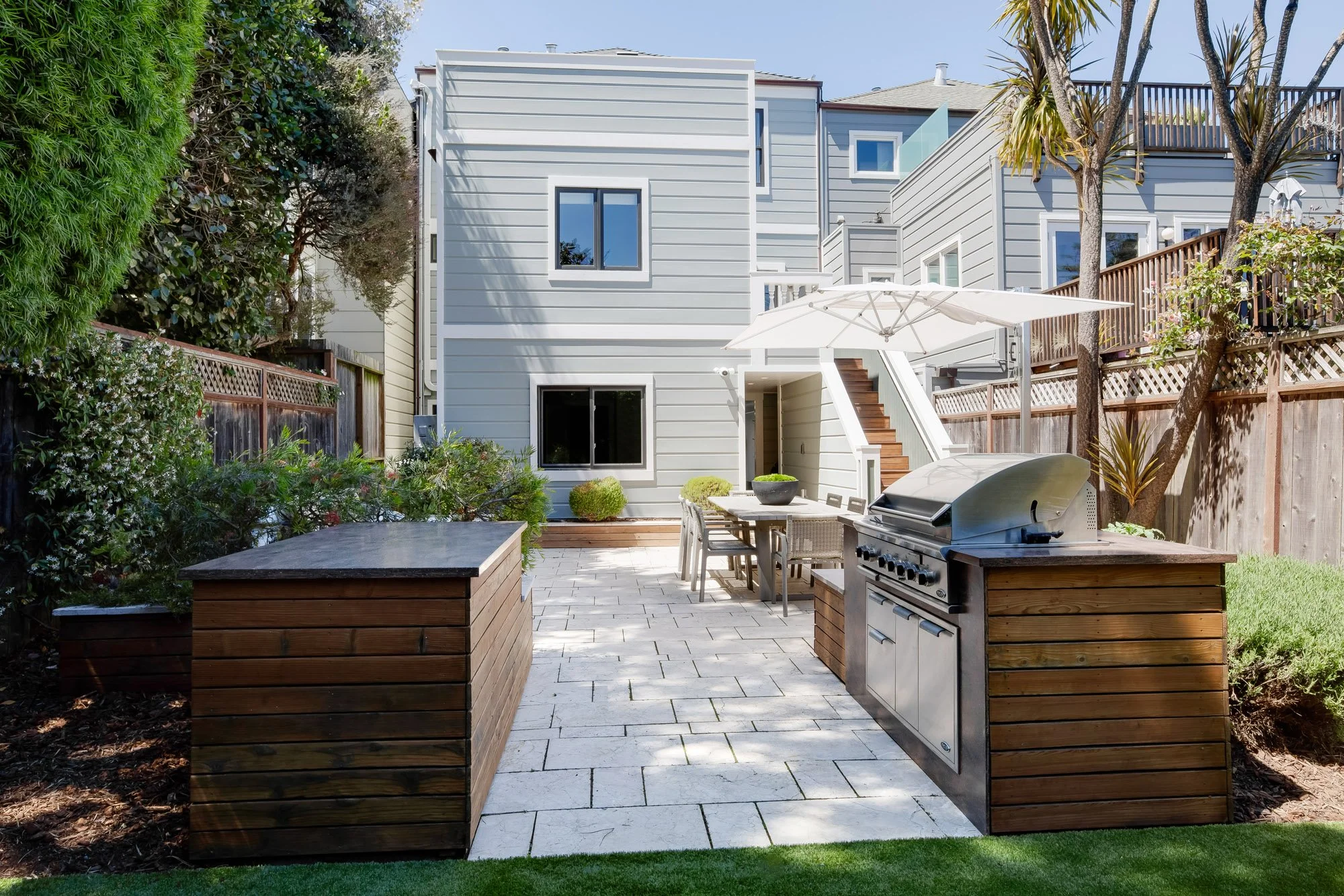 Backyard of 771 Elizabeth Street showing the flat paver patio, built-in outdoor kitchen with stainless grill, raised beds, and the home's rear staircase rising to the upper level.