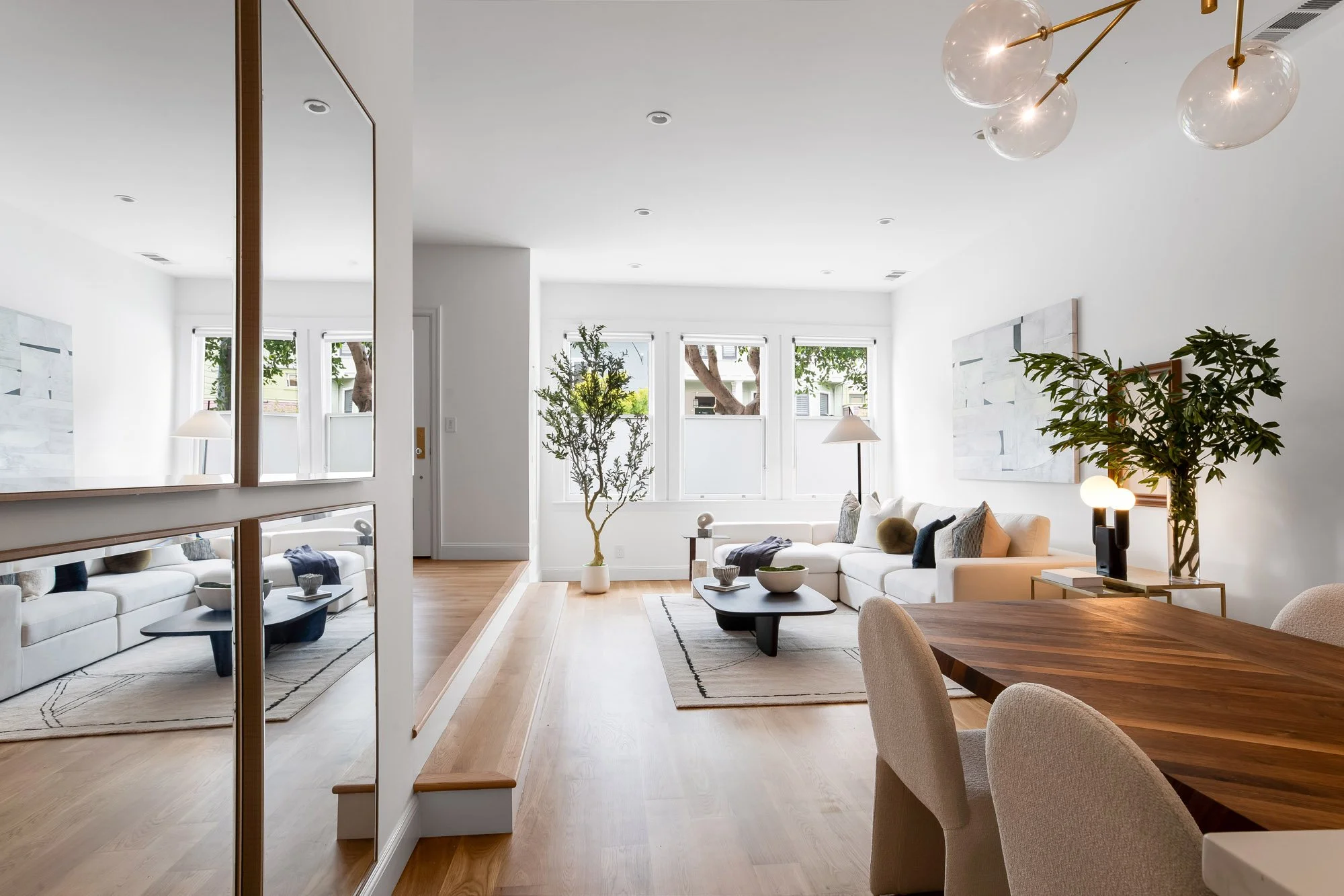 View from dining area into sunlit living room at 4545 18th Street San Francisco TIC featuring framed mirror panels, cream sectional sofa, organic black coffee table, white oak hardwood floors, and gold globe chandelier