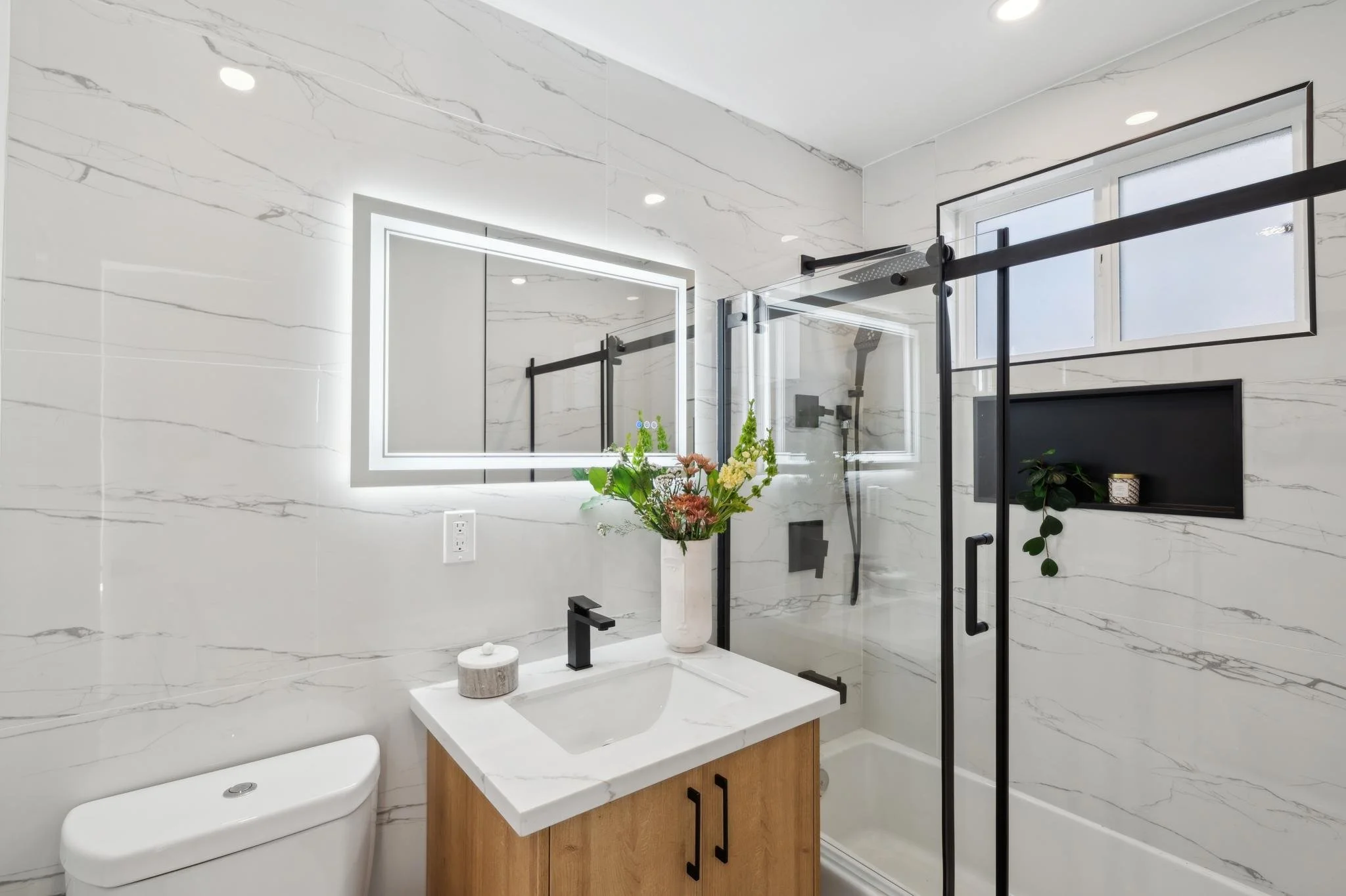 Spa-inspired bathroom with floor-to-ceiling marble tile, a back-lit LED vanity mirror, a modern wood vanity, and a glass-enclosed shower with black trim.