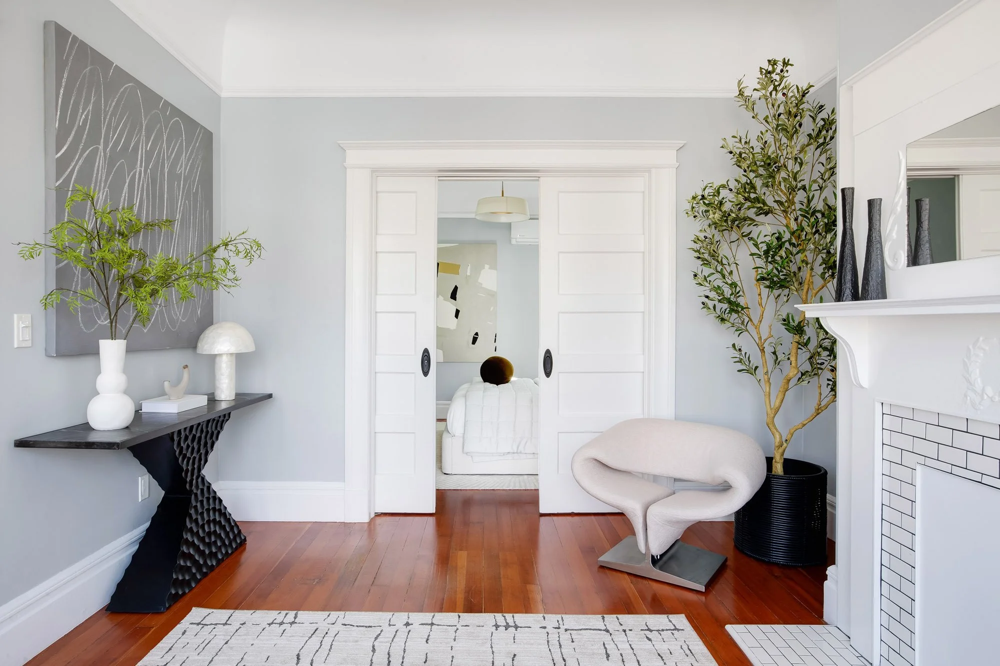 Double pocket doors open between living roo  and bedroom, with an olive tree and fireplace mantel nearby