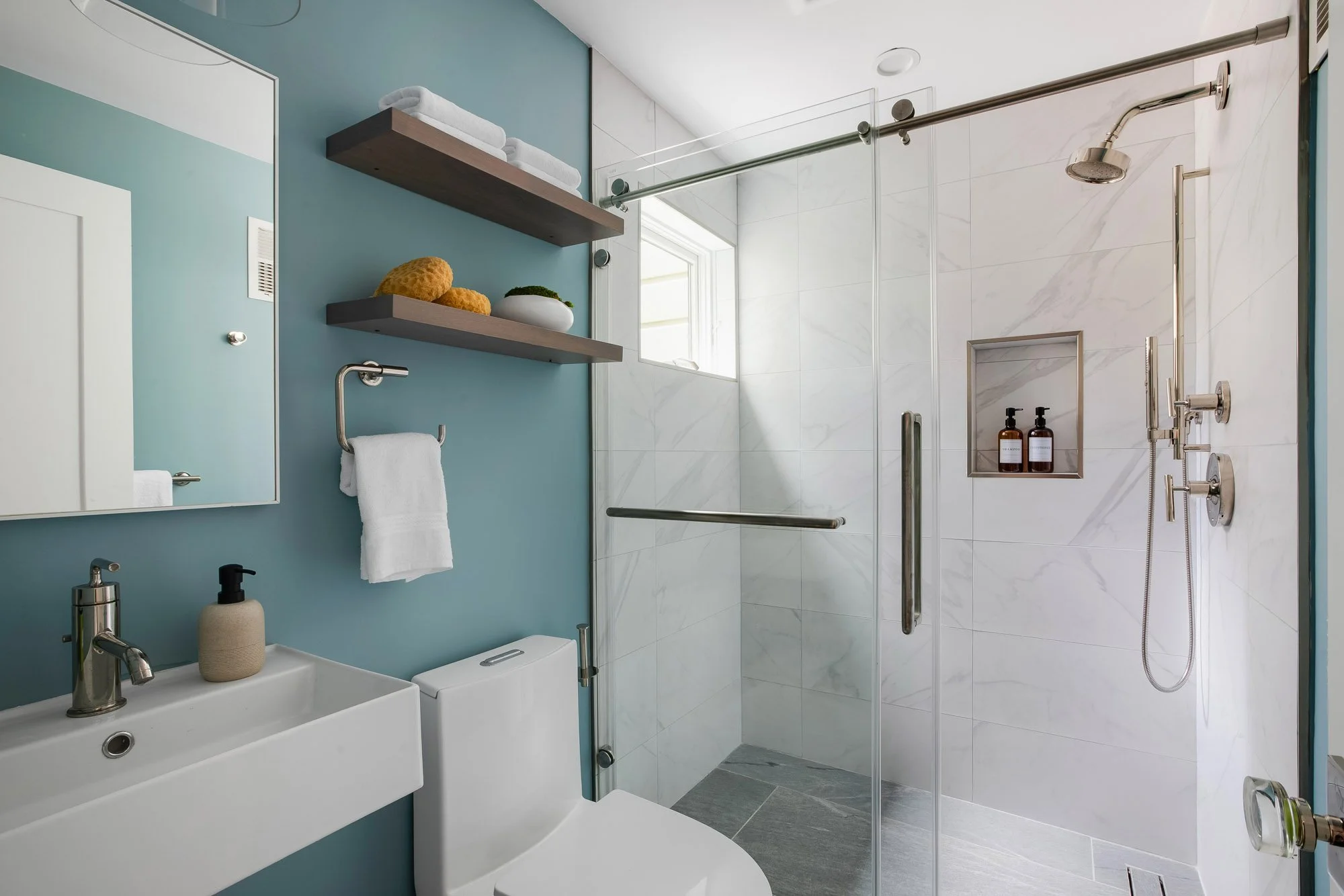 Lower-level bath at 771 Elizabeth Street featuring a glass-enclosed marble walk-in shower with built-in niche, floating wood shelves, a wall-mount sink, and a teal wall.