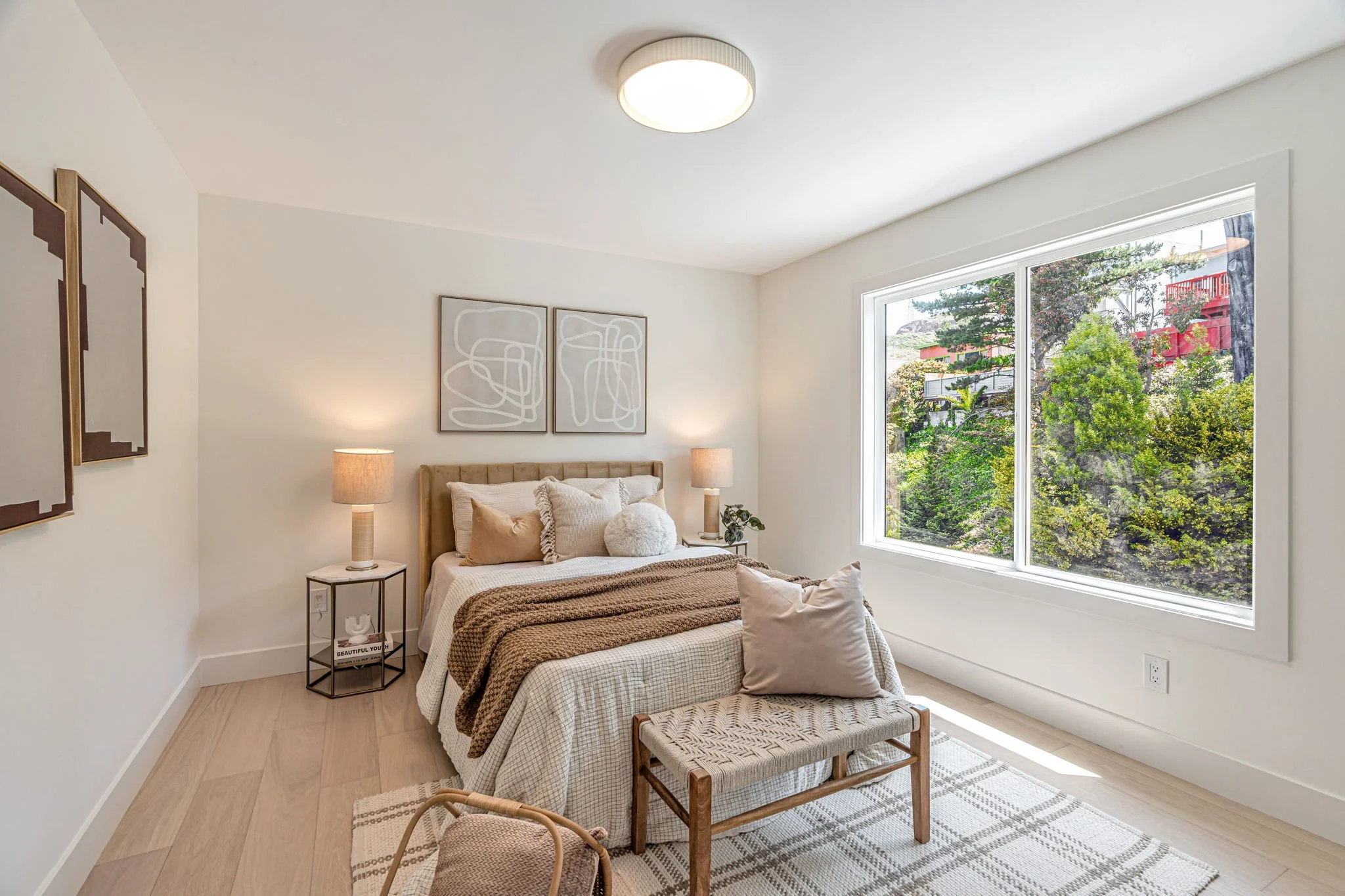A sunny bedroom featuring a large window with a view of hillside greenery. The bed is styled with a tan velvet headboard, neutral bedding, and geometric nightstands with modern lamps.