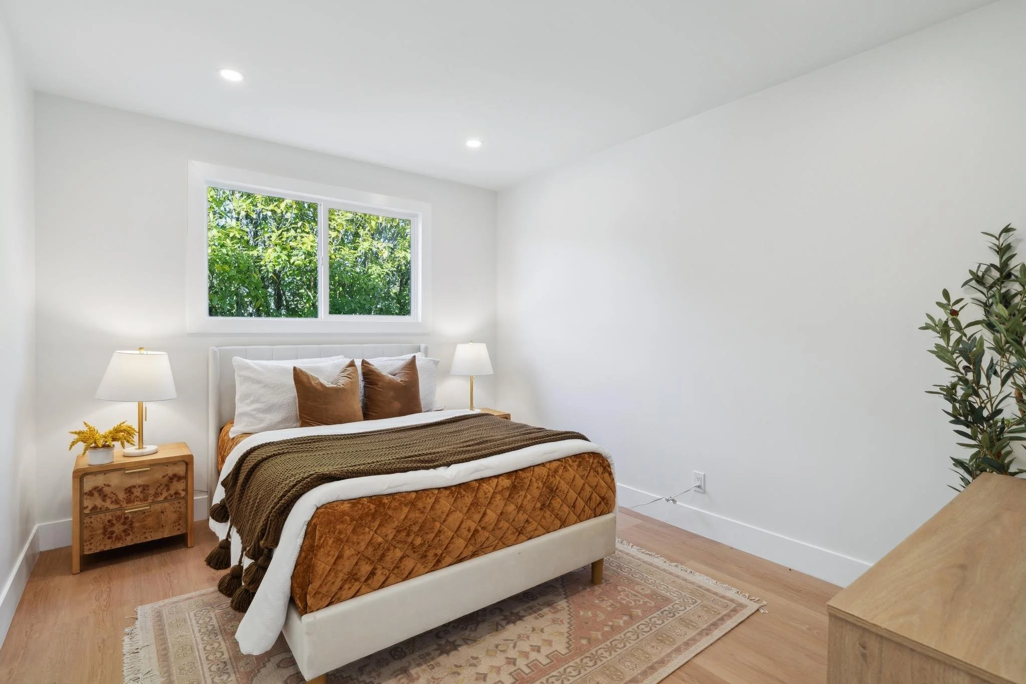 Third guest bedroom featuring warm earth tones, a velvet quilted bedspread, and a large window overlooking lush garden foliage.