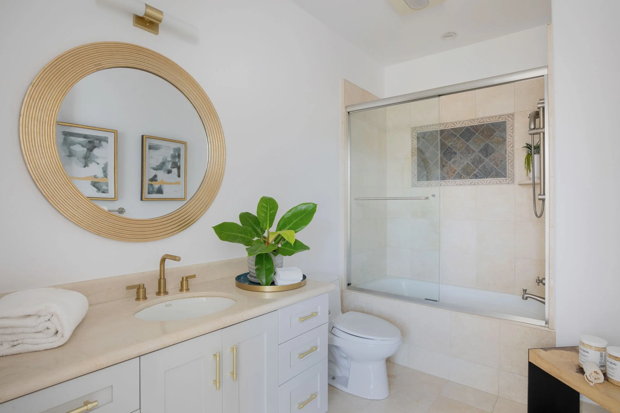 Modern guest bathroom featuring a white vanity with gold hardware, a round wood-framed mirror, and a neutral tile bathtub surround with decorative mosaic accents.