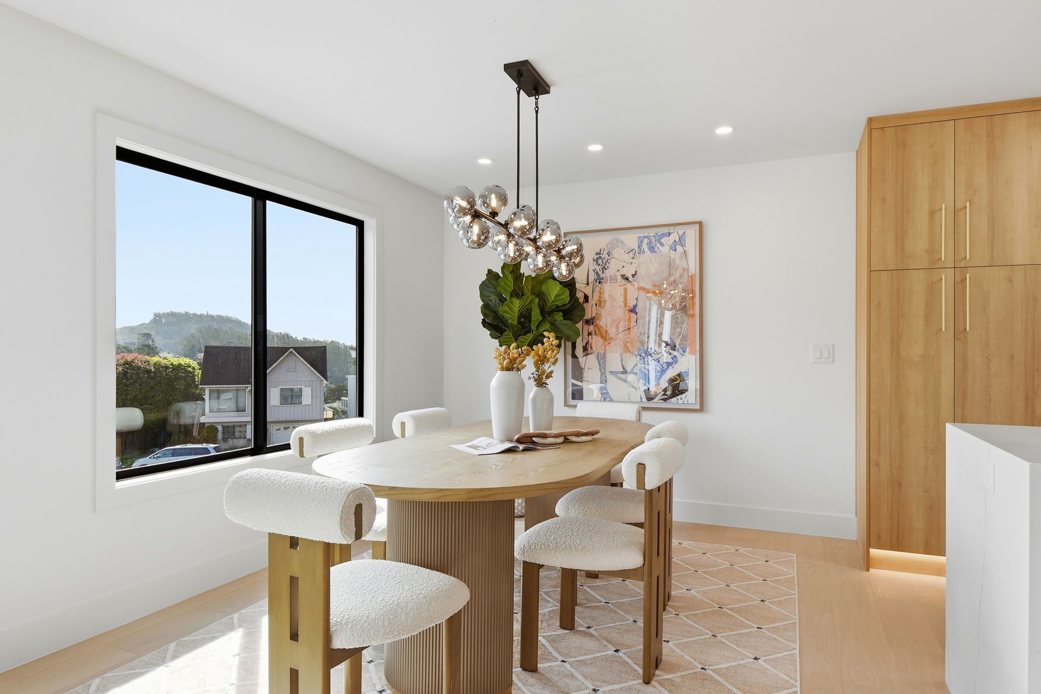 Elegant formal dining room with an oval wood table, modern orb lighting, and a large window overlooking the lush hills of San Francisco.