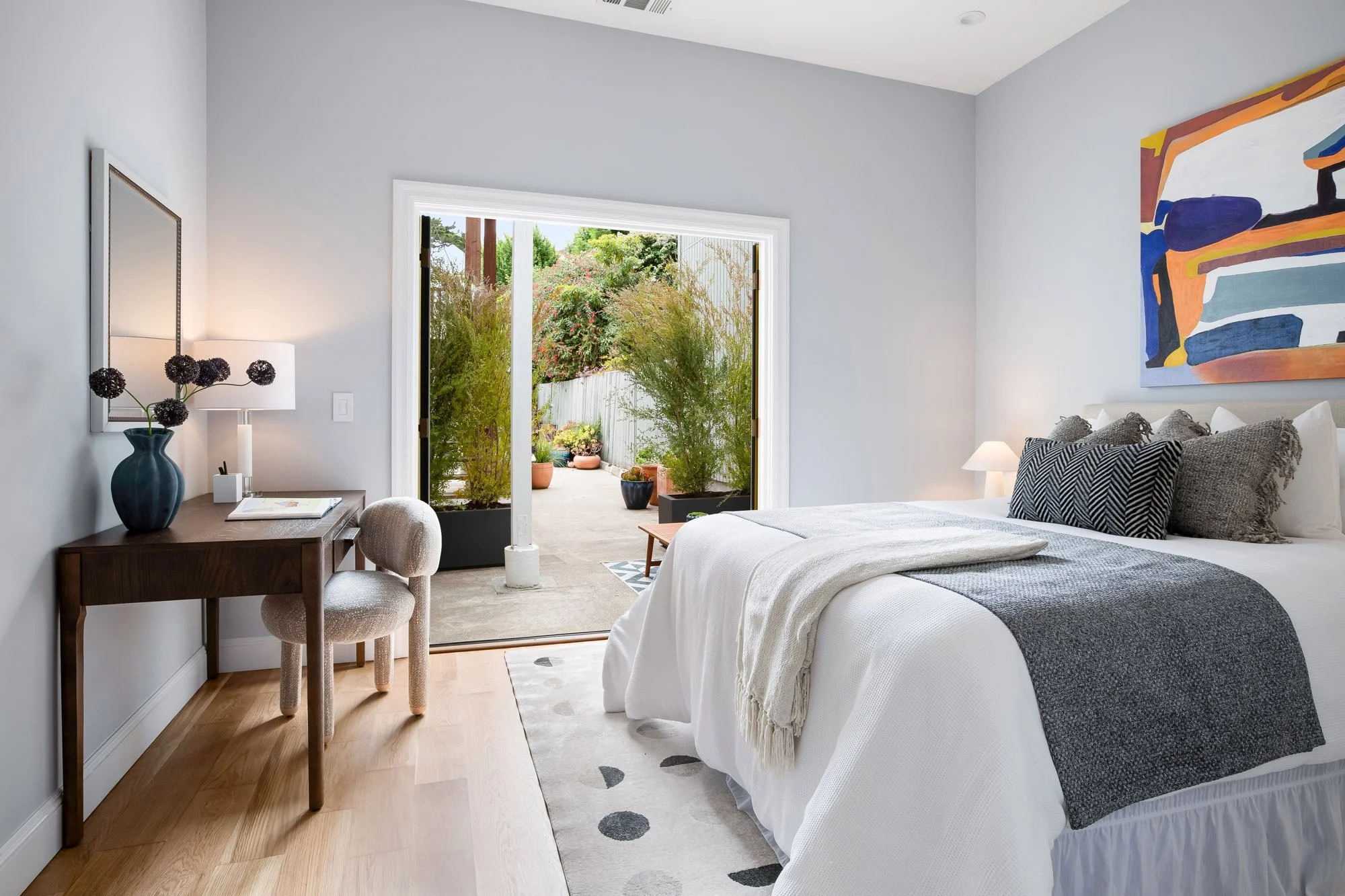 Second bedroom in 4545 18th Street San Francisco TIC with upholstered platform bed, walnut desk, bouclé accent chair, large-scale colorful abstract painting, and open bifold glass doors leading to private landscaped garden patio