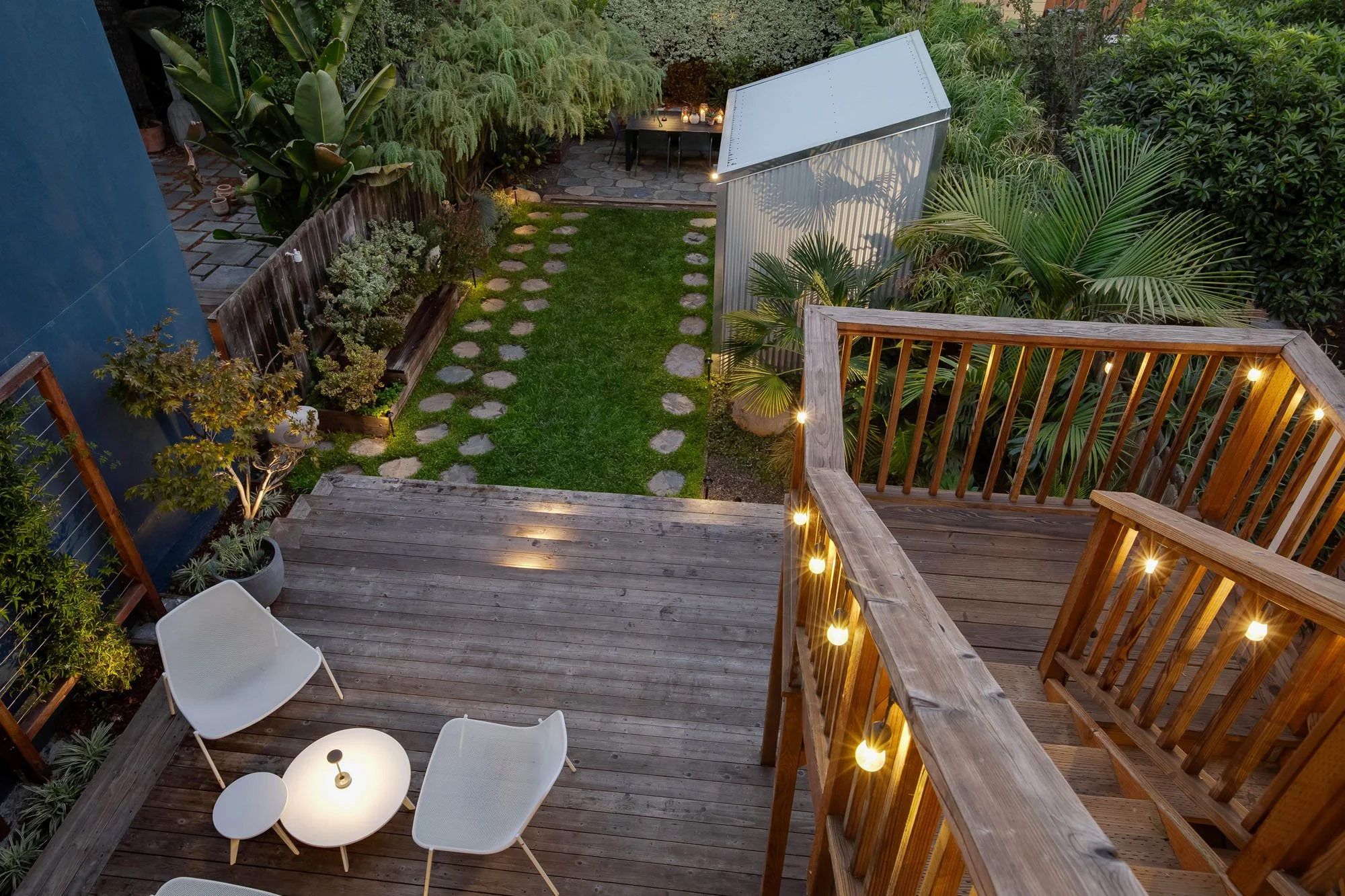 Multi-level backyard at 246 Detroit Street, SF, featuring a tiered wooden entertaining deck with string lights, modern white outdoor lounge furniture, a green lawn, and a corrugated metal garden studio.